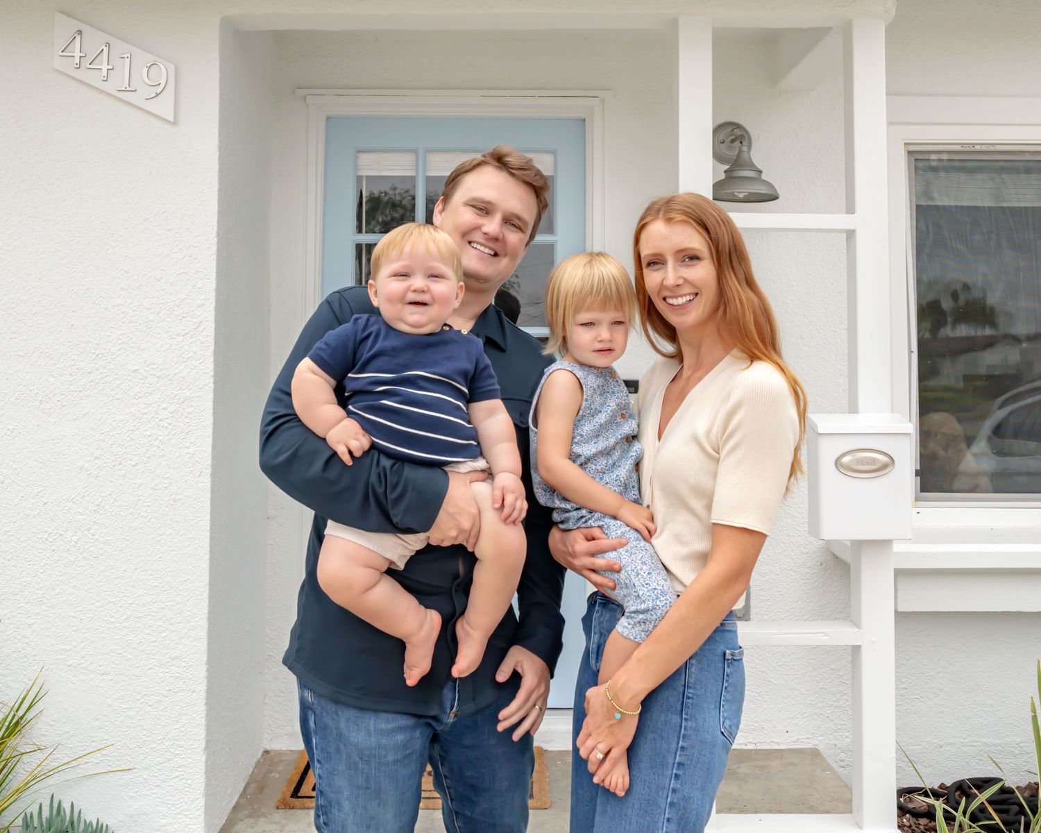 Dad holds a very chunky infant and mom holds their toddler, standing on their front porch smiling at the camera.  Doughman Photography
