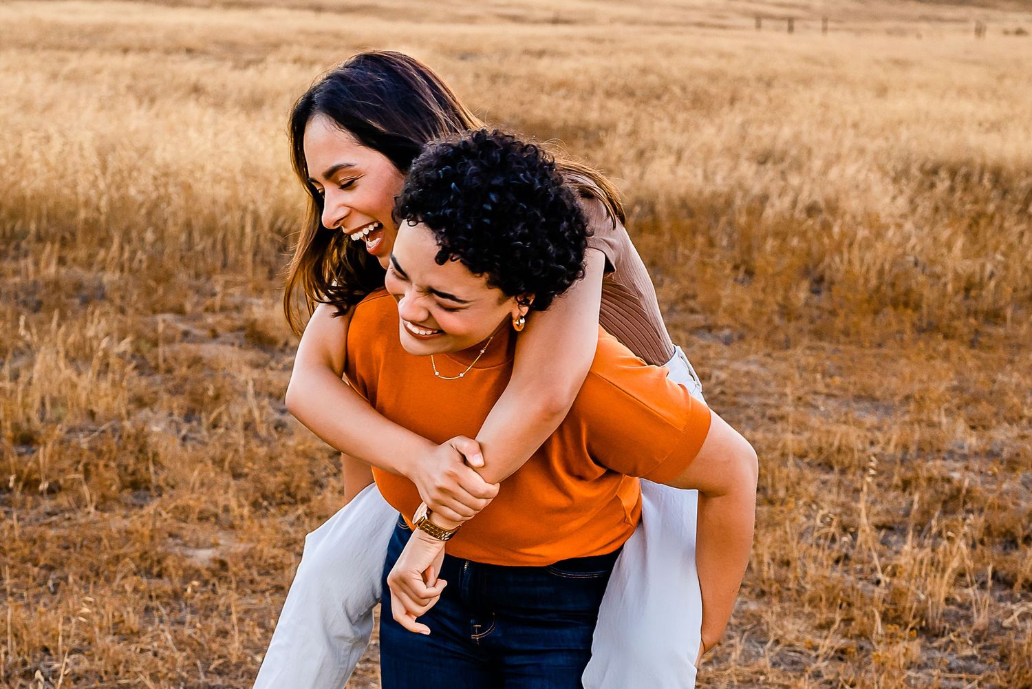 Best friend pippy back ride in open field.One friend is wearing a deep orange sweater. The other an Amber colored tee.Warm & Inviting colors
