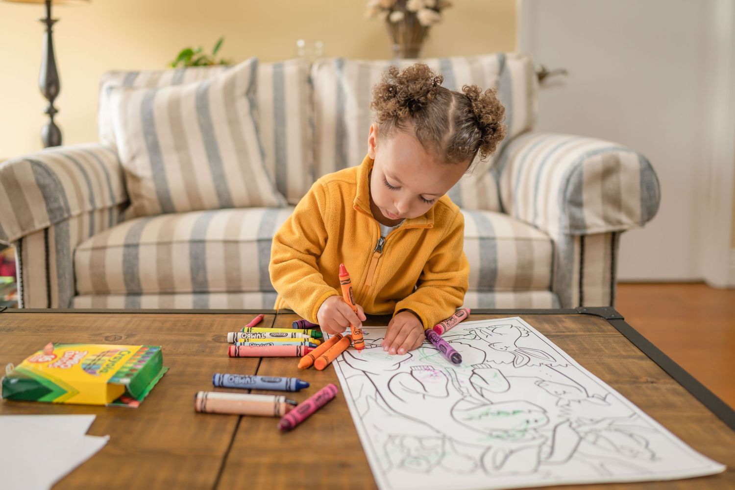 Toddler holding a jumbo crayon upside down as she colors a dinosaur picture on the coffee table. Other crayons are spilled out on the table.