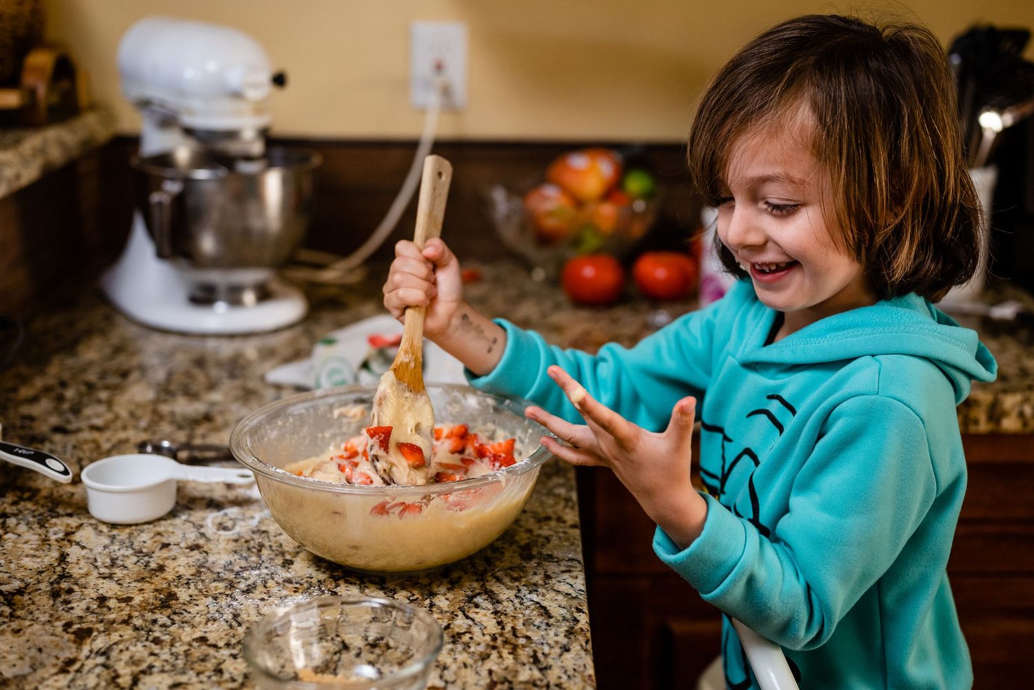 Young child mixing muffin batter looks at the batter spilled on his hand with a laugh.  Doughman Photography.
