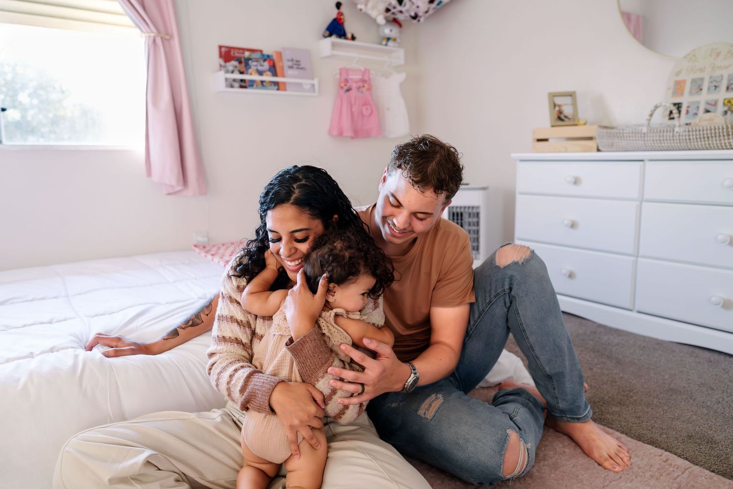 Mom and dad sitting on the floor next to the bed. Toddler gives mom a big loving hug. Mom wraps her in a warm embrace. Dad leans in to hug.