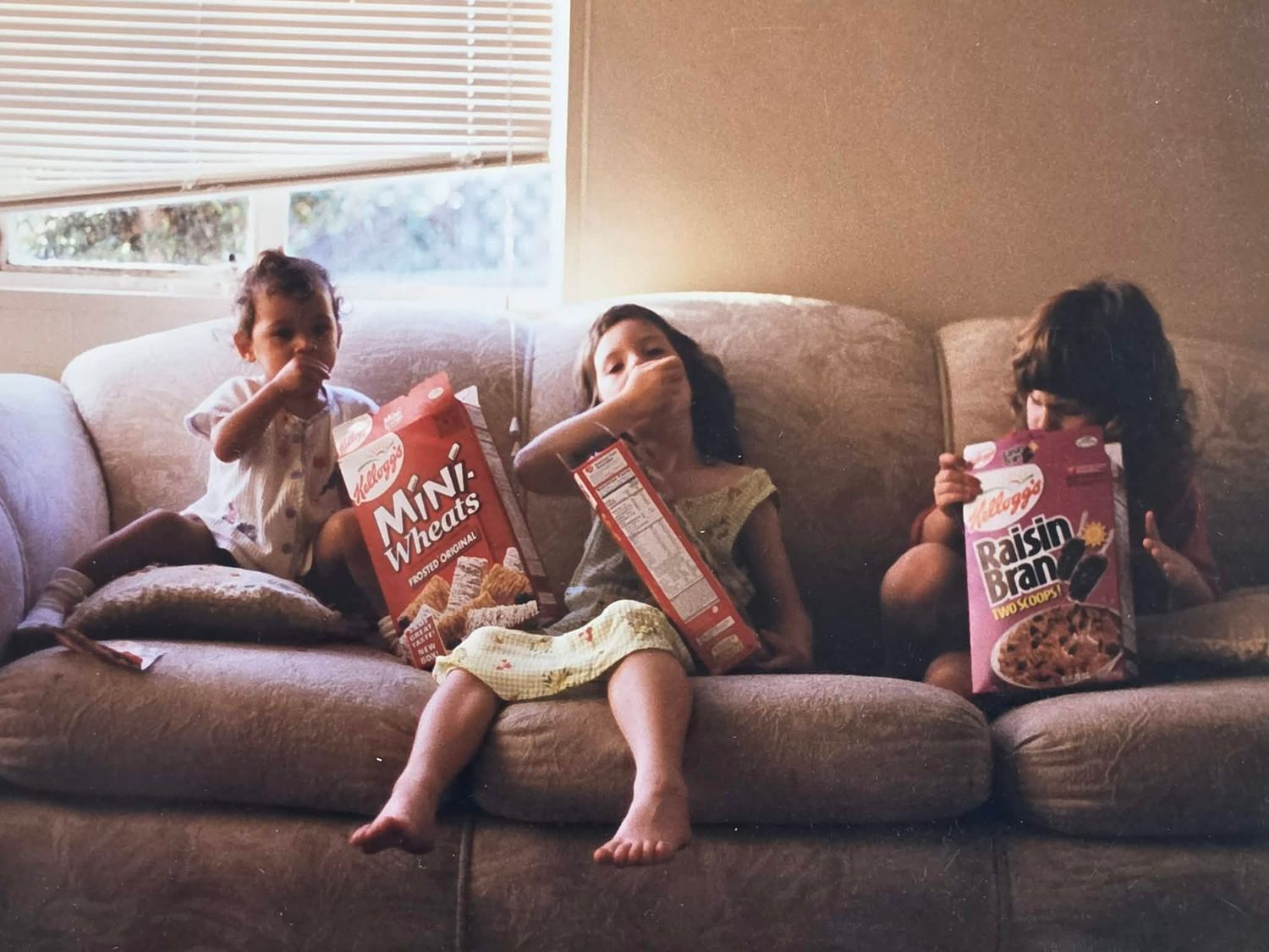 Three siblings eating cereal together on a couch at home