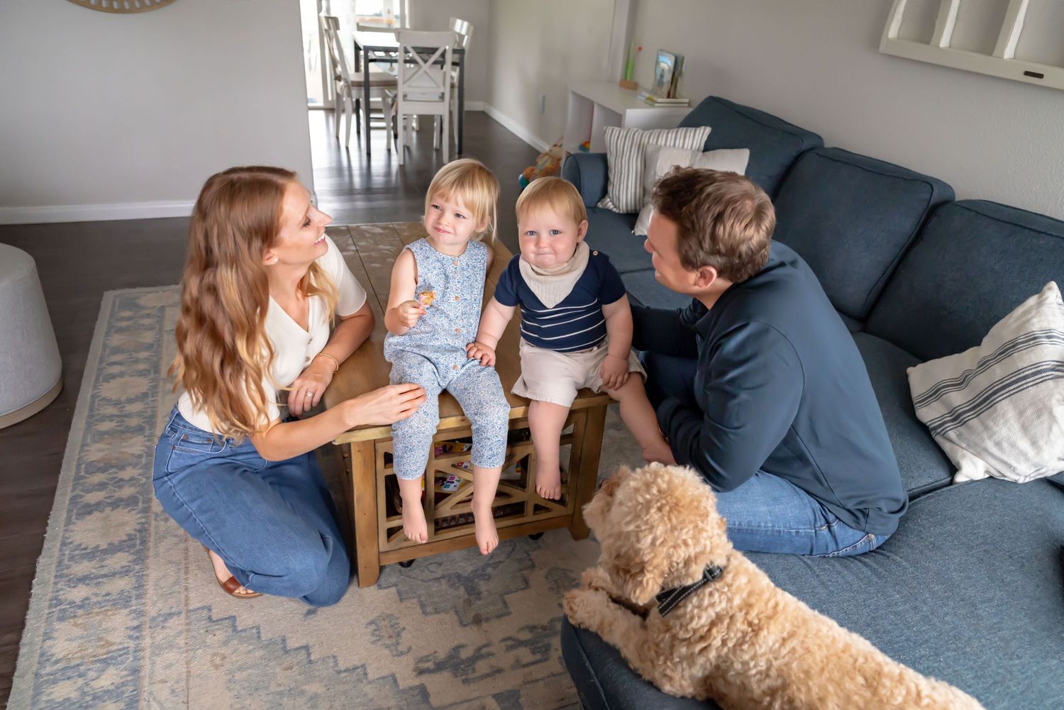 Slight overhead of two toddlers sitting on the coffee table looking at the camera, mom and dad are on either side looking at the kids.