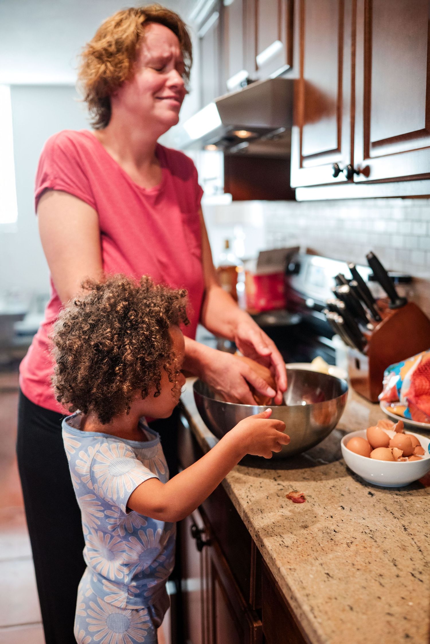 Mom tosses head back in laughter as daughter finishes cracking eggs. Doughman Photography.