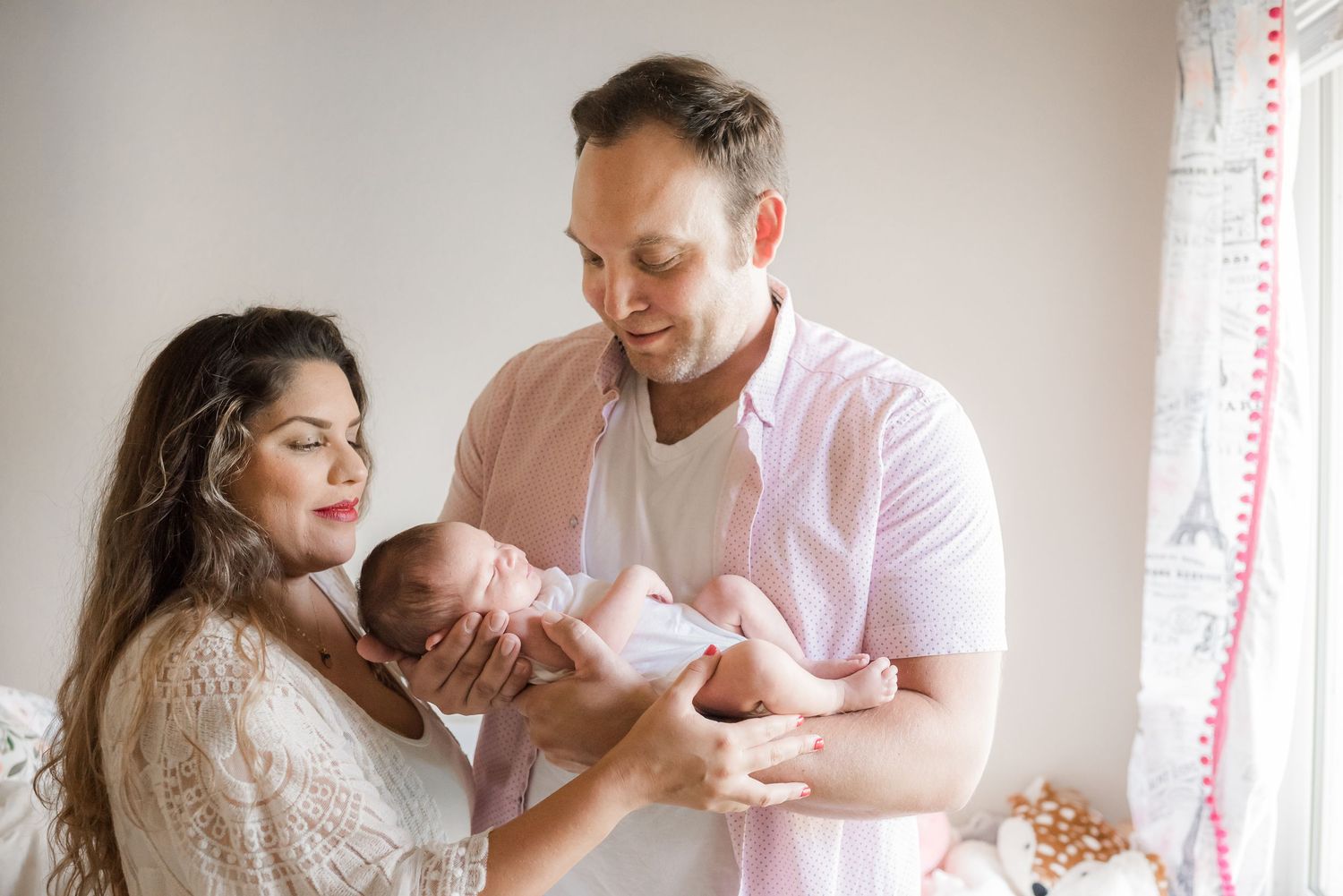 Dad standing in nursery holding newborn daughter. Mom gives them gentle embrace. In-home newborn photography. Doughman Photography