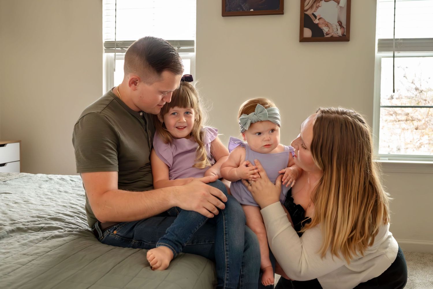 Family of four in bedroom. Toddler smiles, leans into dad sitting on edge of bed while mom sits on the floor holding baby. Doughman Photo