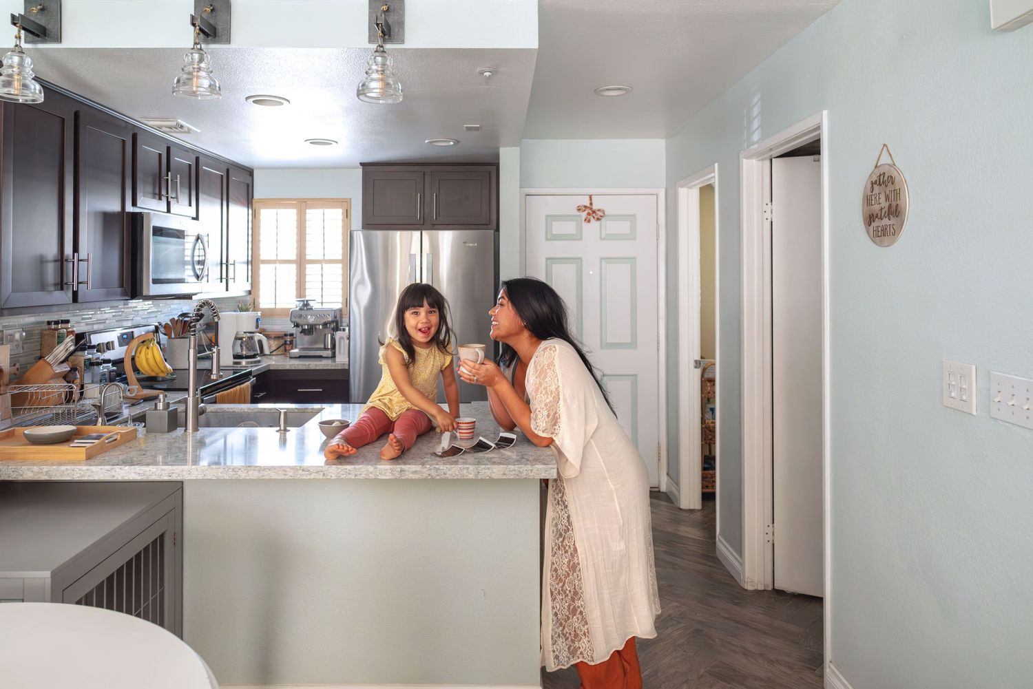 Toddler sits on kitchen counter; mom leans in  holding a cup of coffee. Both are smiling, enjoying the peaceful moment. Doughman Photography
