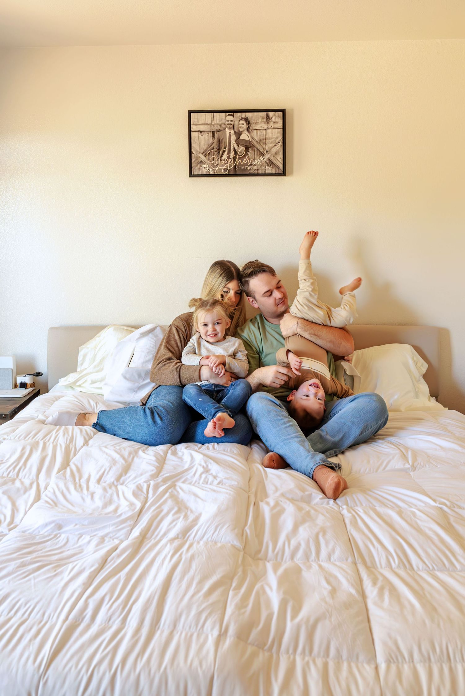 Family sitting close together on bed. Toddler snuggles with mom. Dad holds younger brother upside down.  Doughman Photography