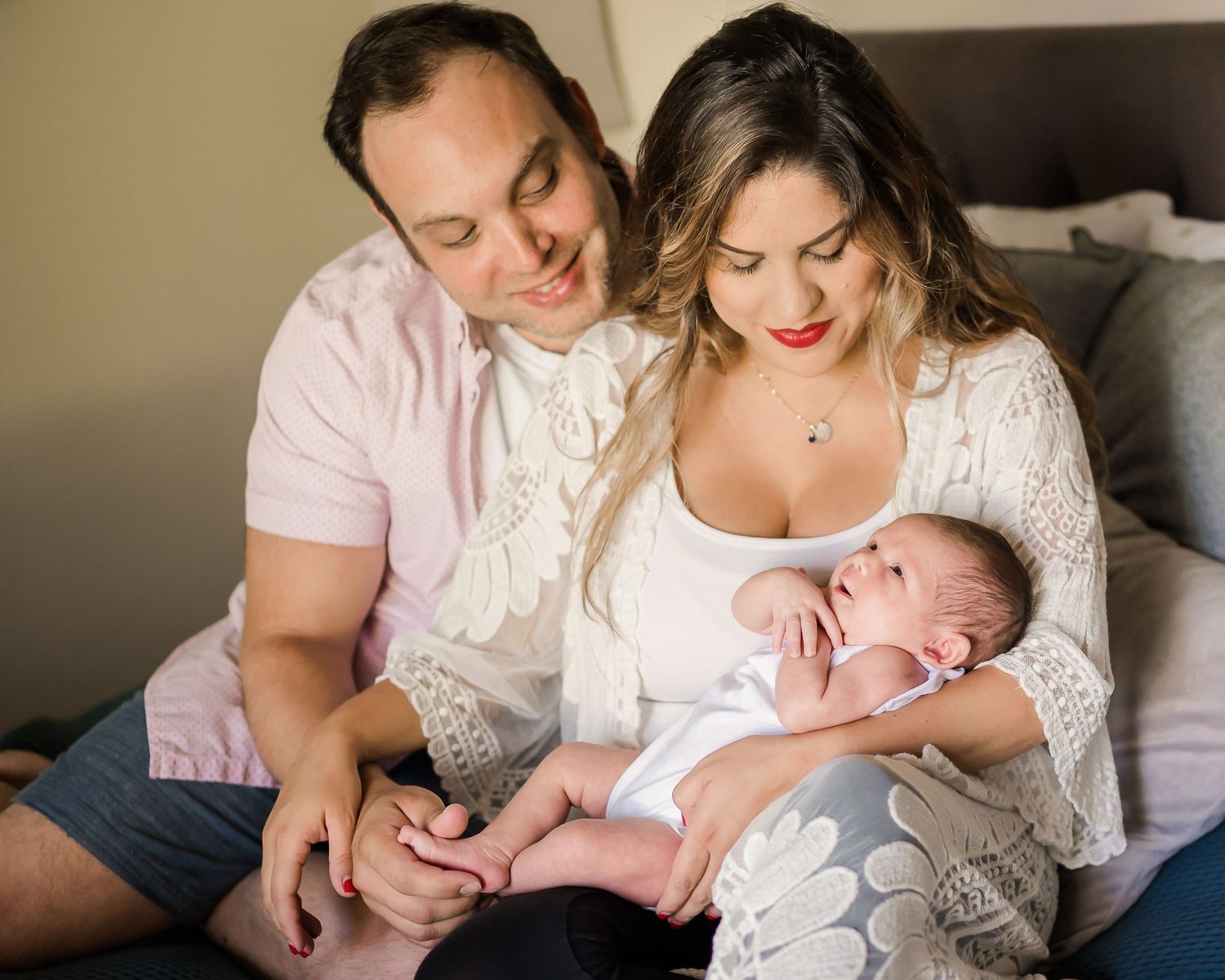 Parents snuggled together on bed, both lovingly looking at their newborn daughter. San Diego In-Home Family Session. Doughman Photography
