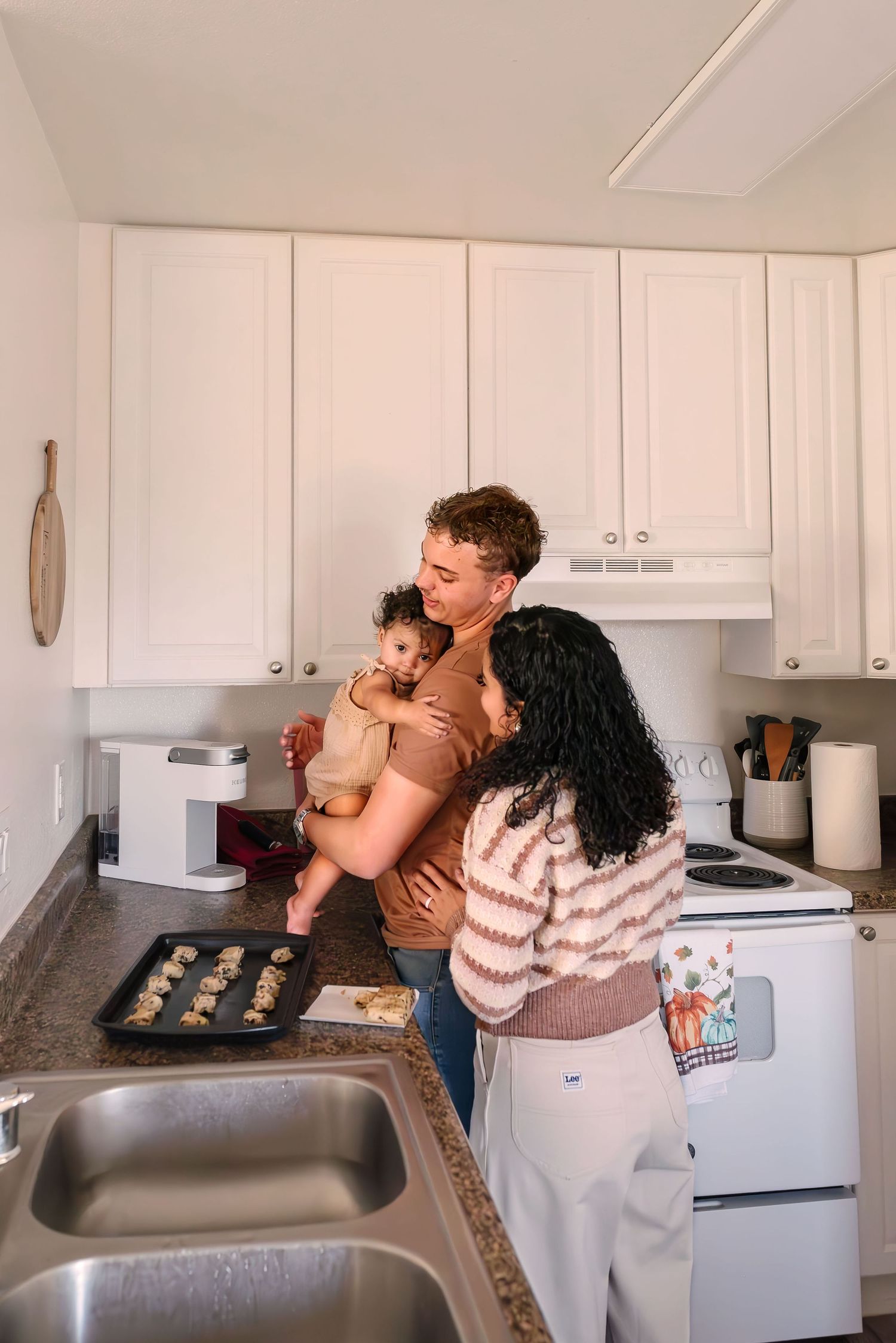 Toddler standing on counter hugging dad. Mom leaning in. Tray of cookies on the counter waiting to go in the oven. Doughman Photography