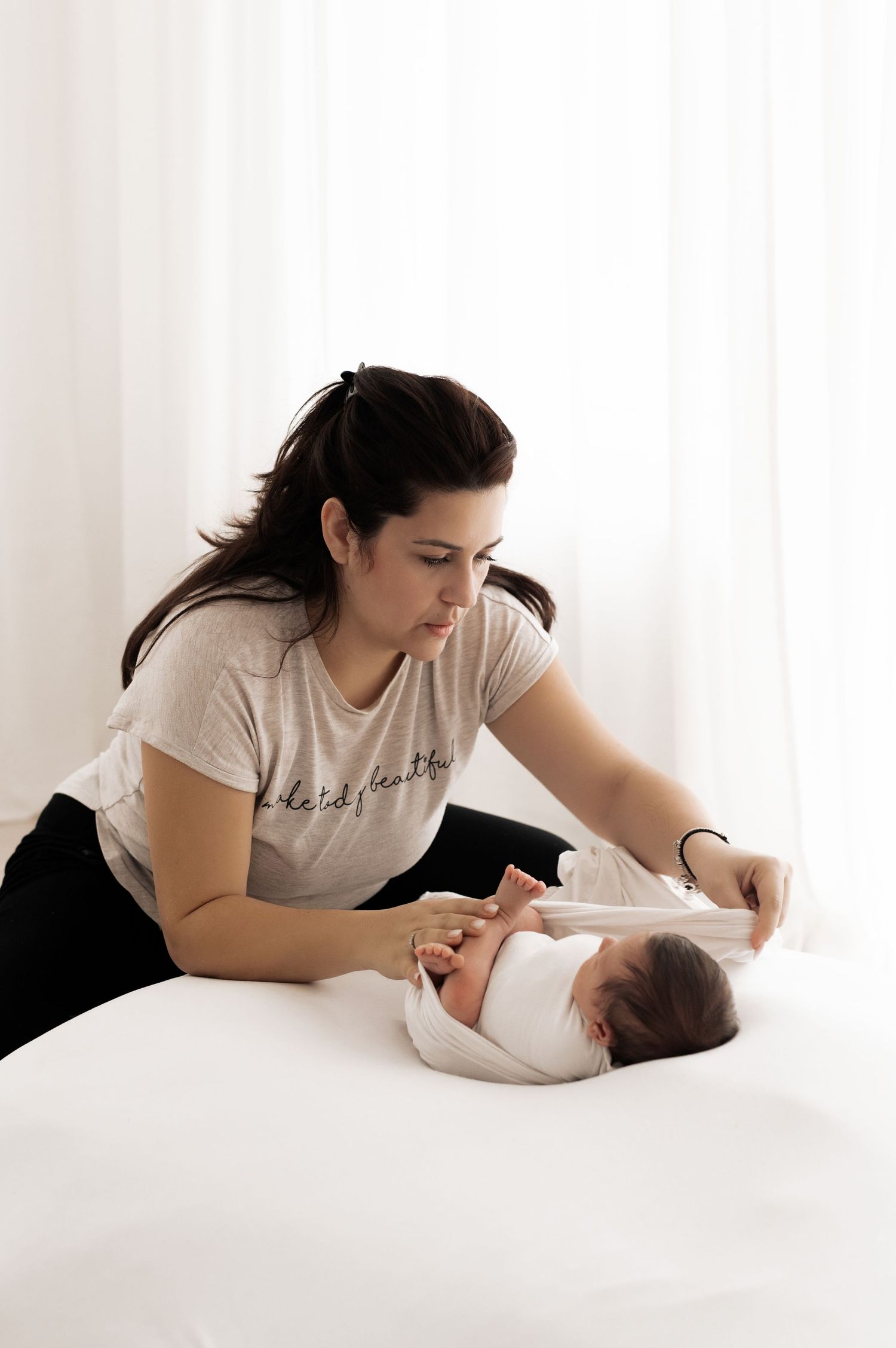 A mother in a casual t-shirt tenderly cares for her baby lying on white bedding in a bright room.