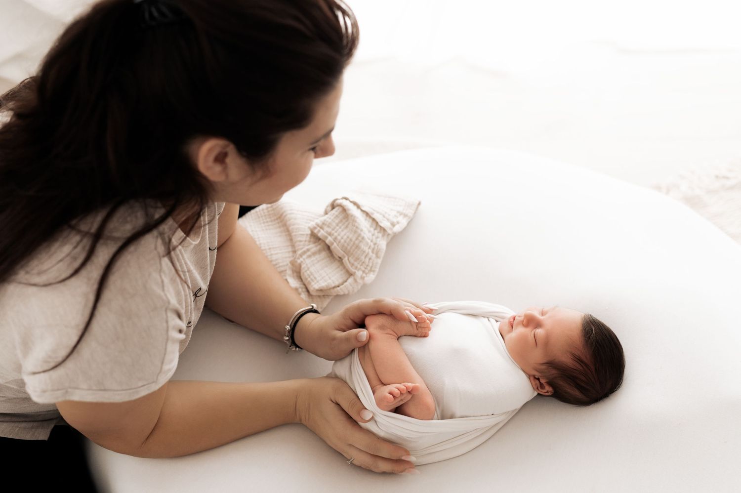 A parent lovingly gazes at their newborn baby while lying together on white bedding.