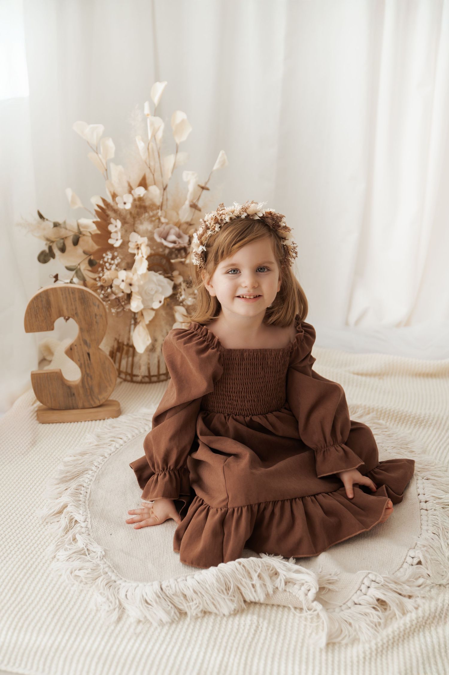 A baby in a brown dress sits on a white textured blanket with dried floral decorations in the background.