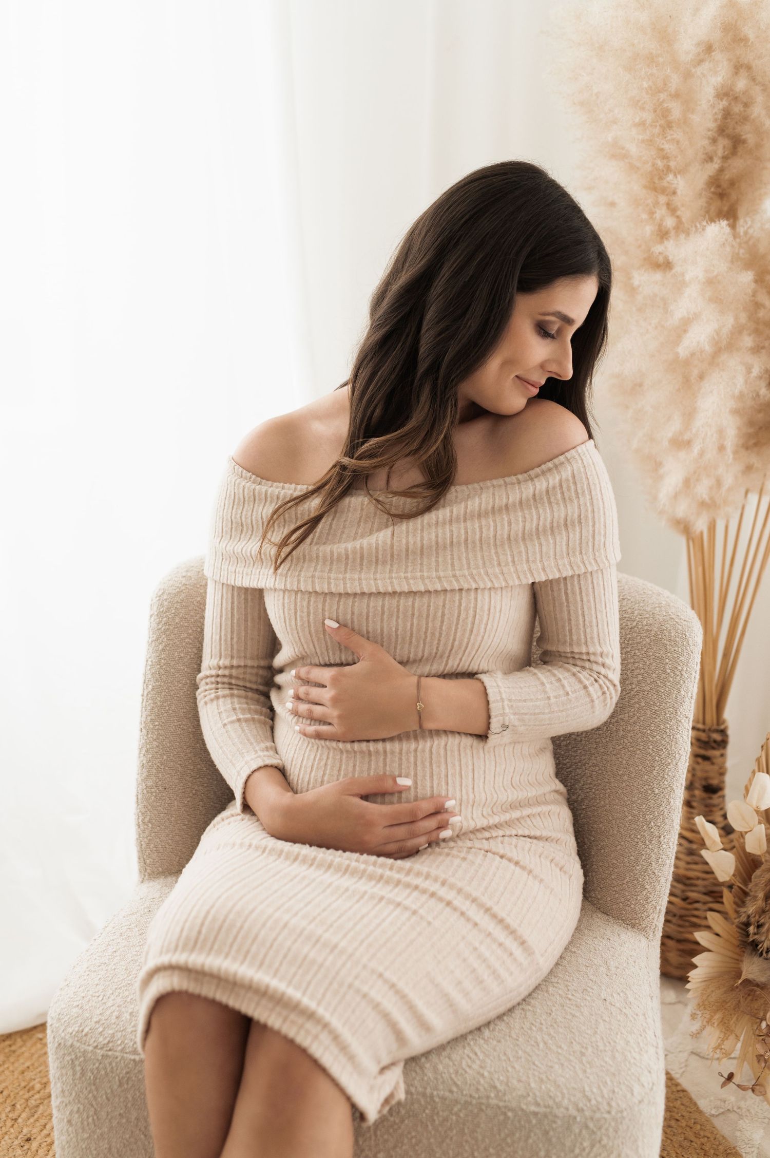 Pregnant woman in beige sweater dress sits on cream armchair with dried pampas grass decor in soft natural lighting.