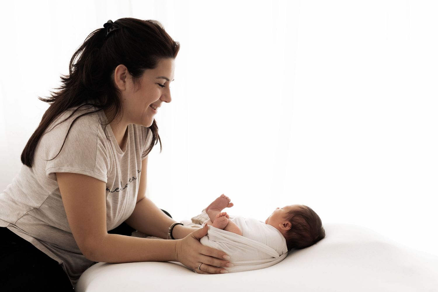 A mother lies on a white bed holding and gazing lovingly at her newborn baby.