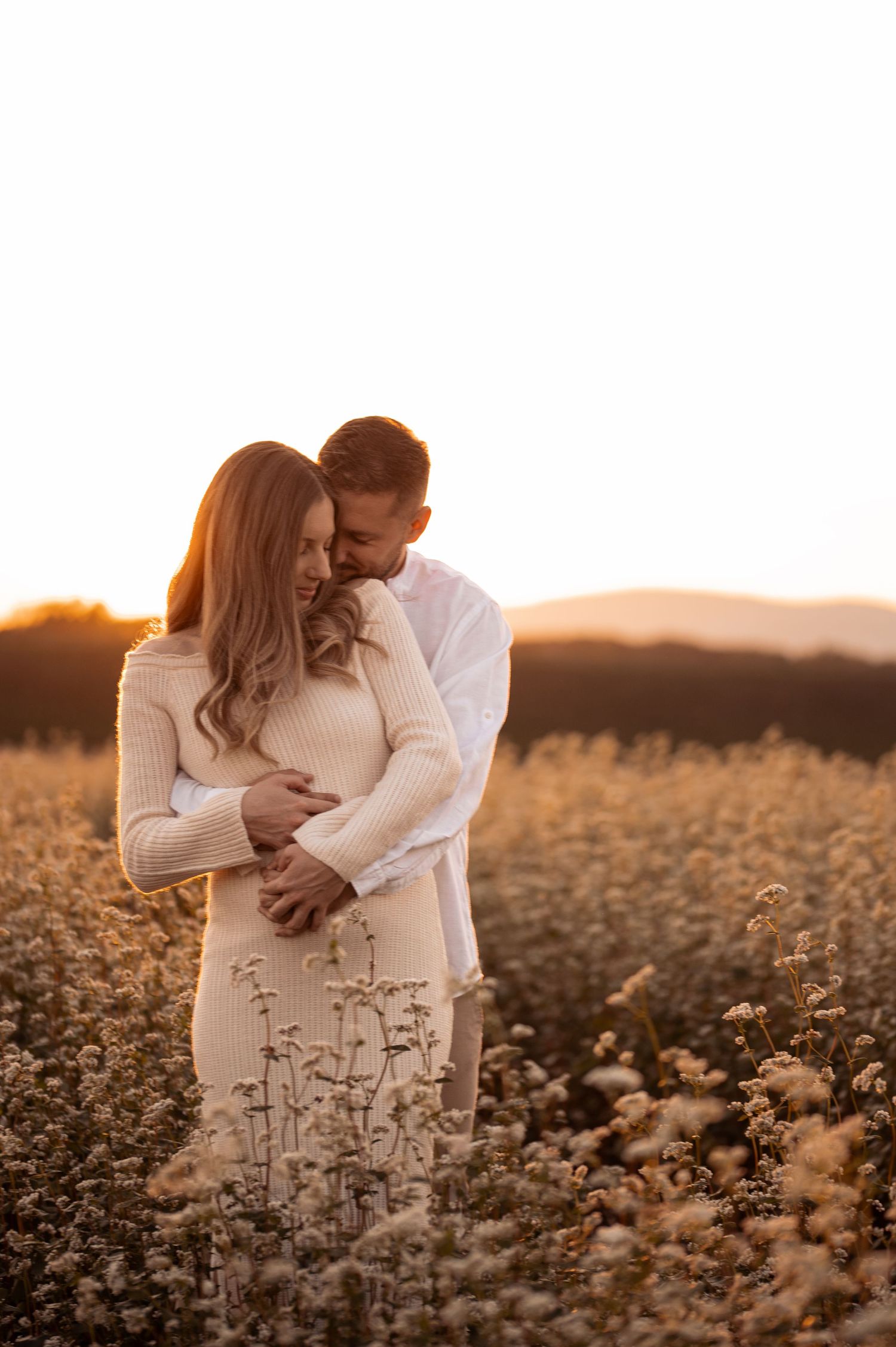 A couple embraces tenderly in a golden field of wildflowers at sunset with warm glowing light.