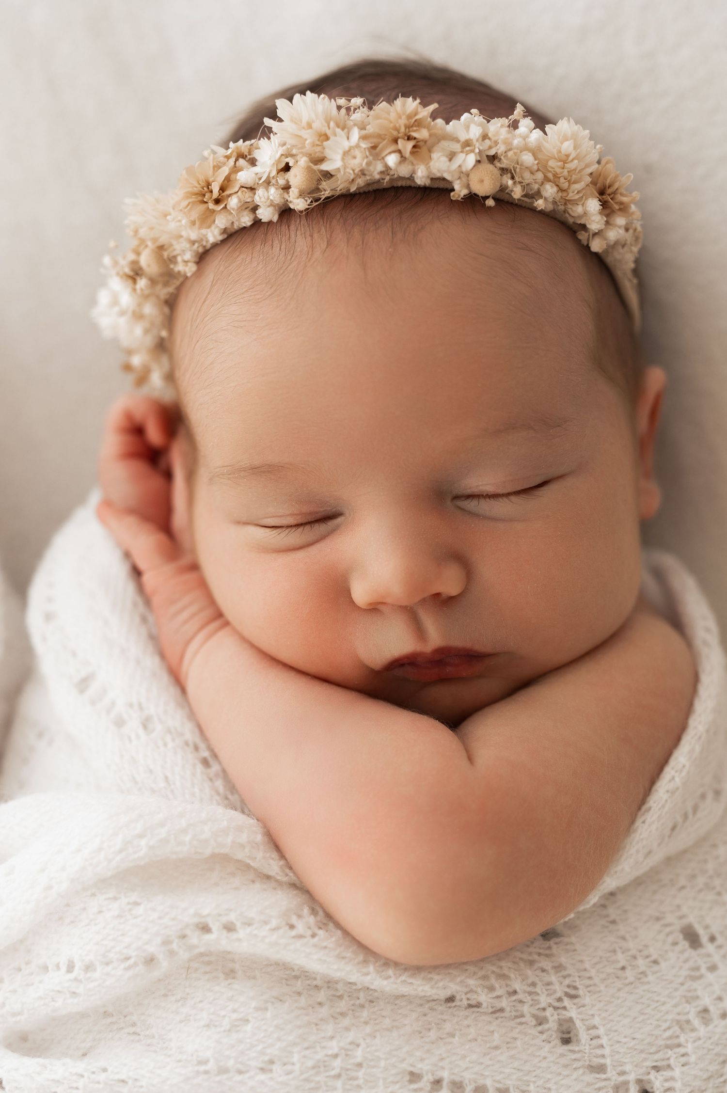 Sleeping newborn baby wearing delicate white lace wrap and floral crown in peaceful studio portrait series.
