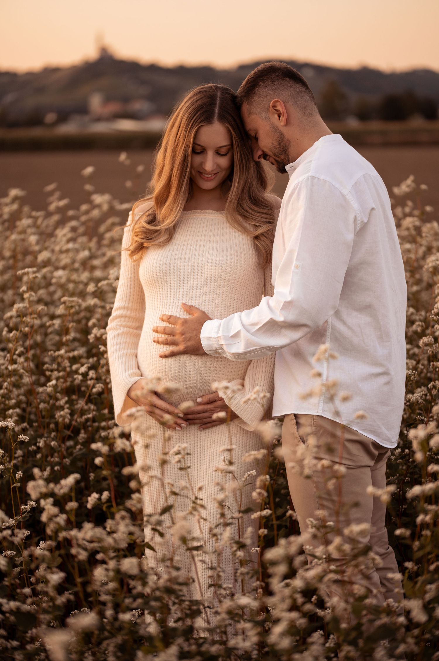 A couple shares an intimate moment in a wildflower field at sunset as they celebrate their upcoming arrival.