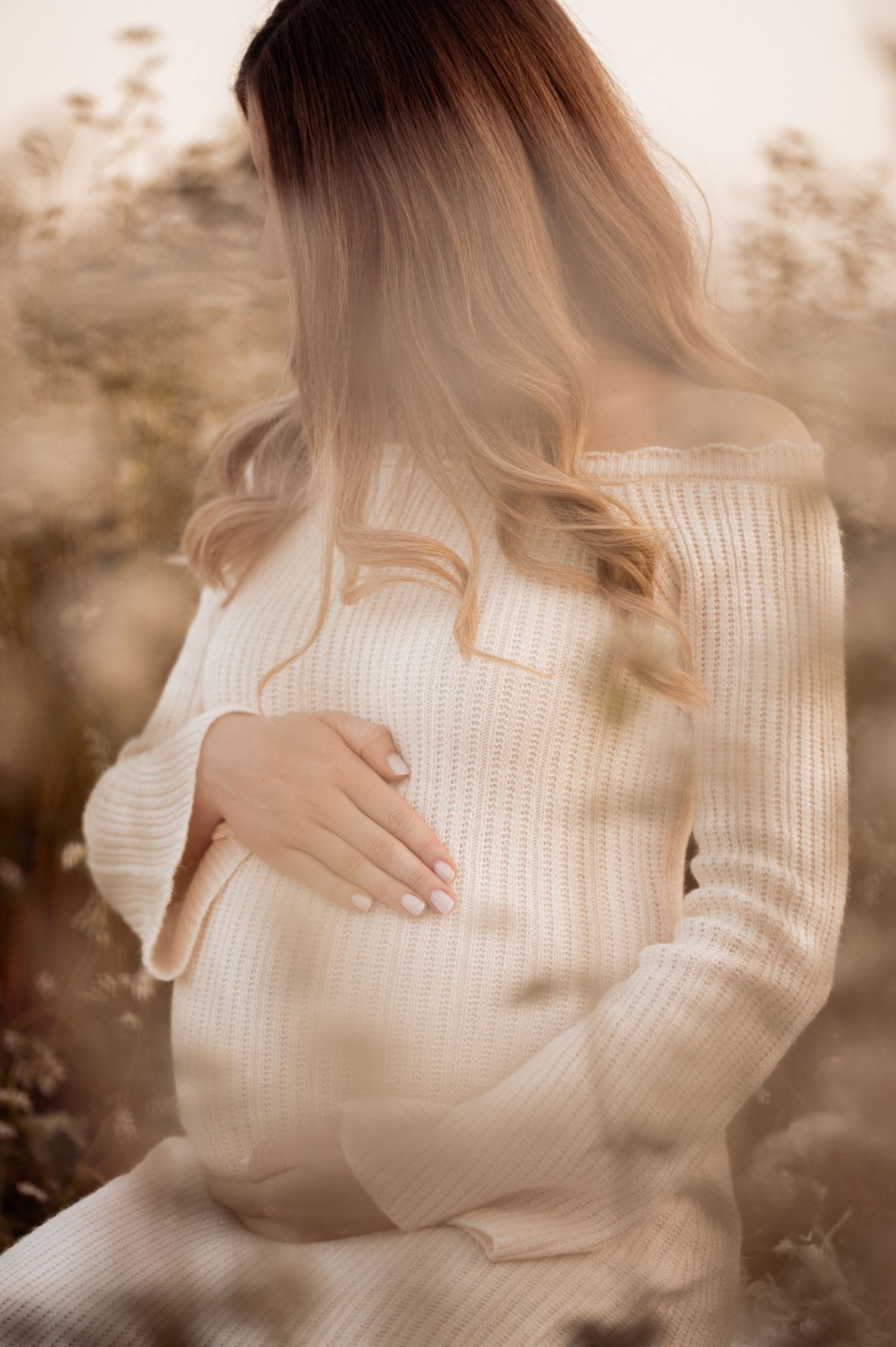 A pregnant woman in a white knit sweater gently cradles her baby bump in a soft, sepia-toned outdoor portrait.