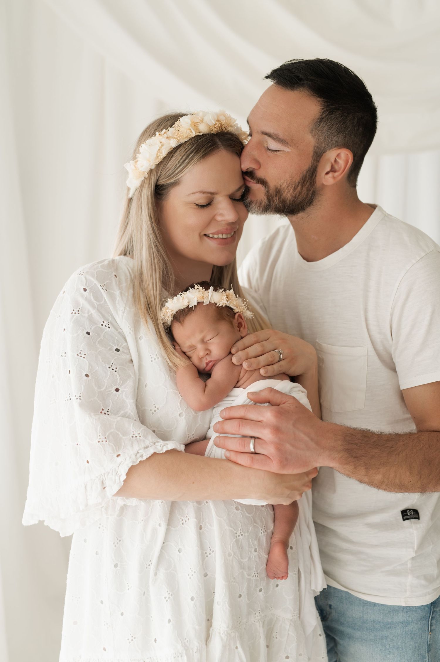 Tender family moment as parents embrace sleeping newborn baby in white nursery setting.