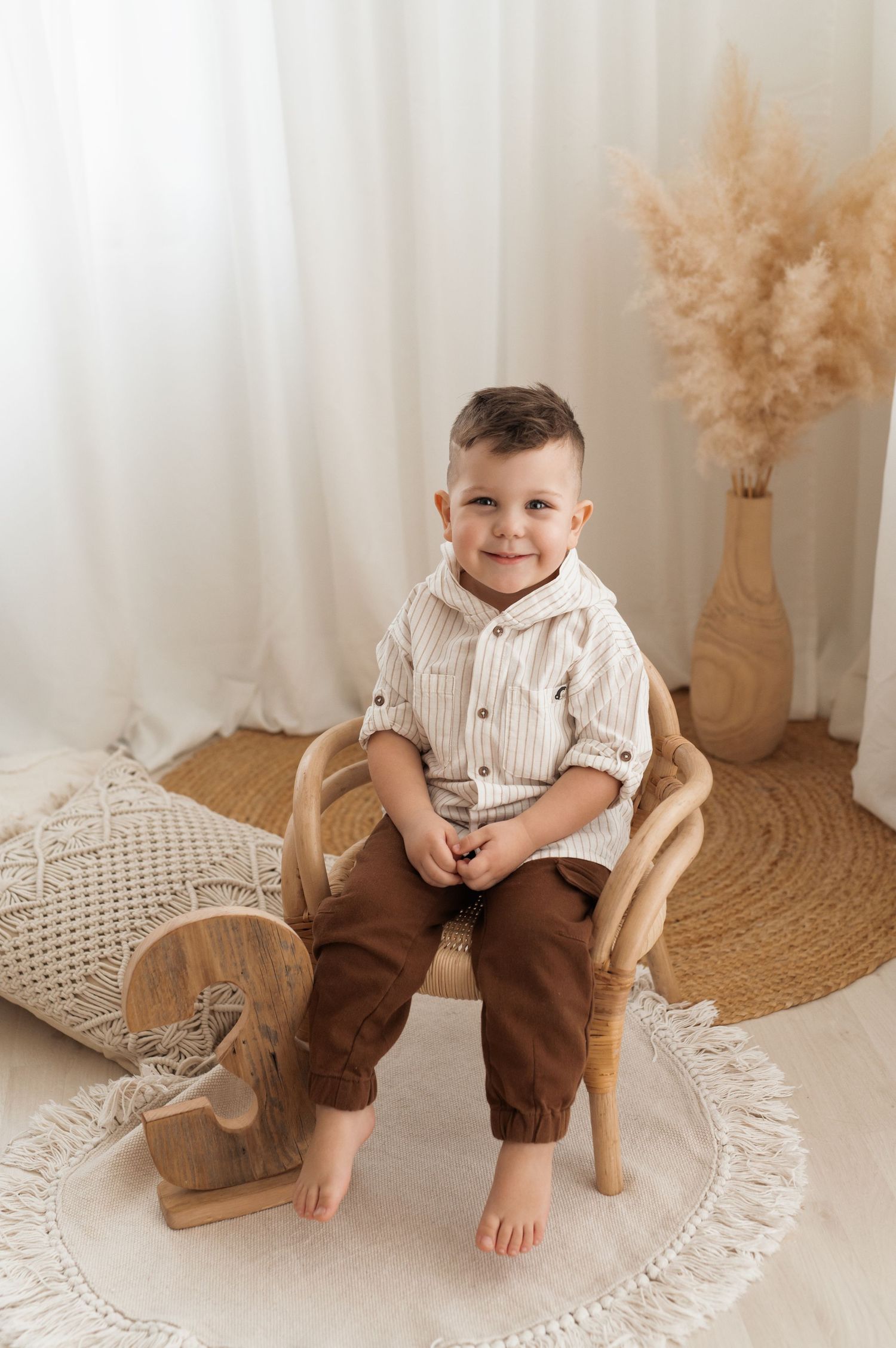 A young child sits barefoot on a wooden chair with pampas grass decor and boho elements in a natural light setting.