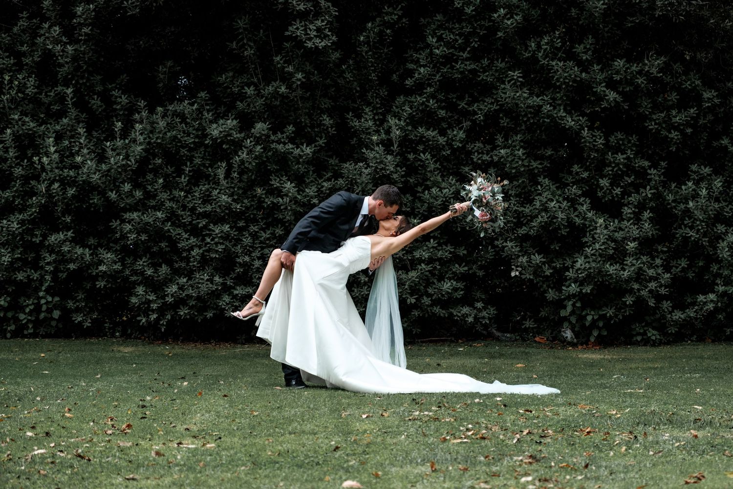A bride in a flowing white dress lays gracefully on green grass surrounded by autumn leaves in a park setting.