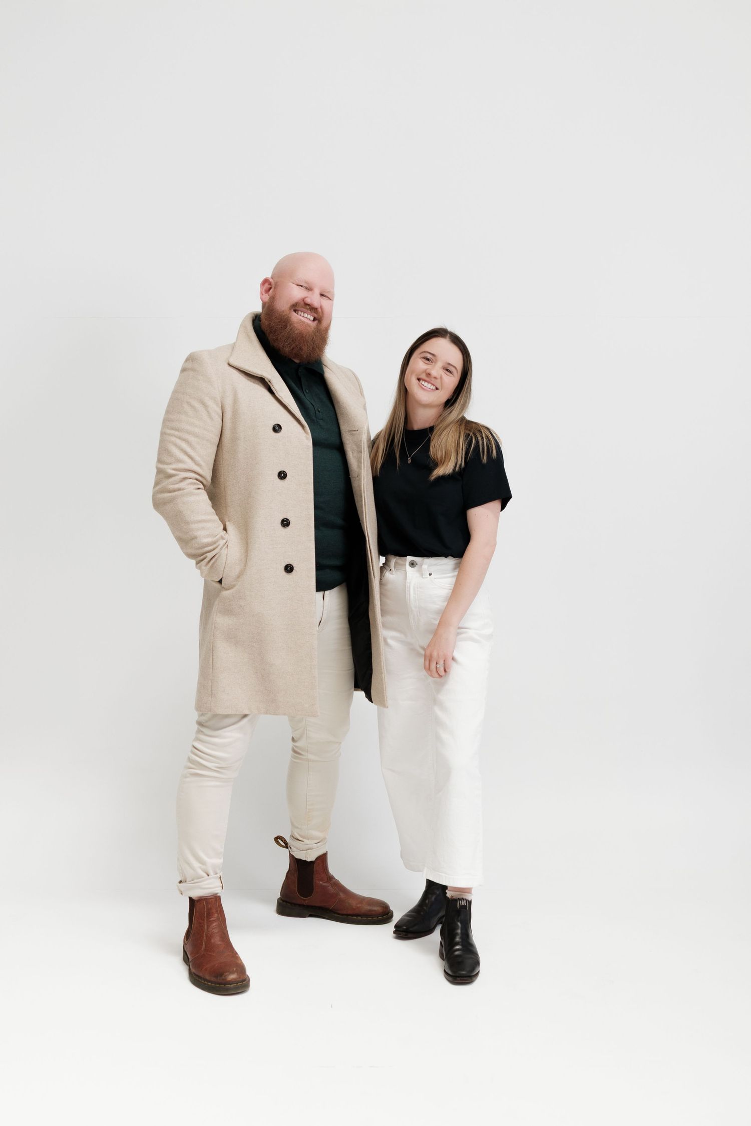 A bearded man in a beige coat and a woman in a black top and white jeans smile together against a white background.