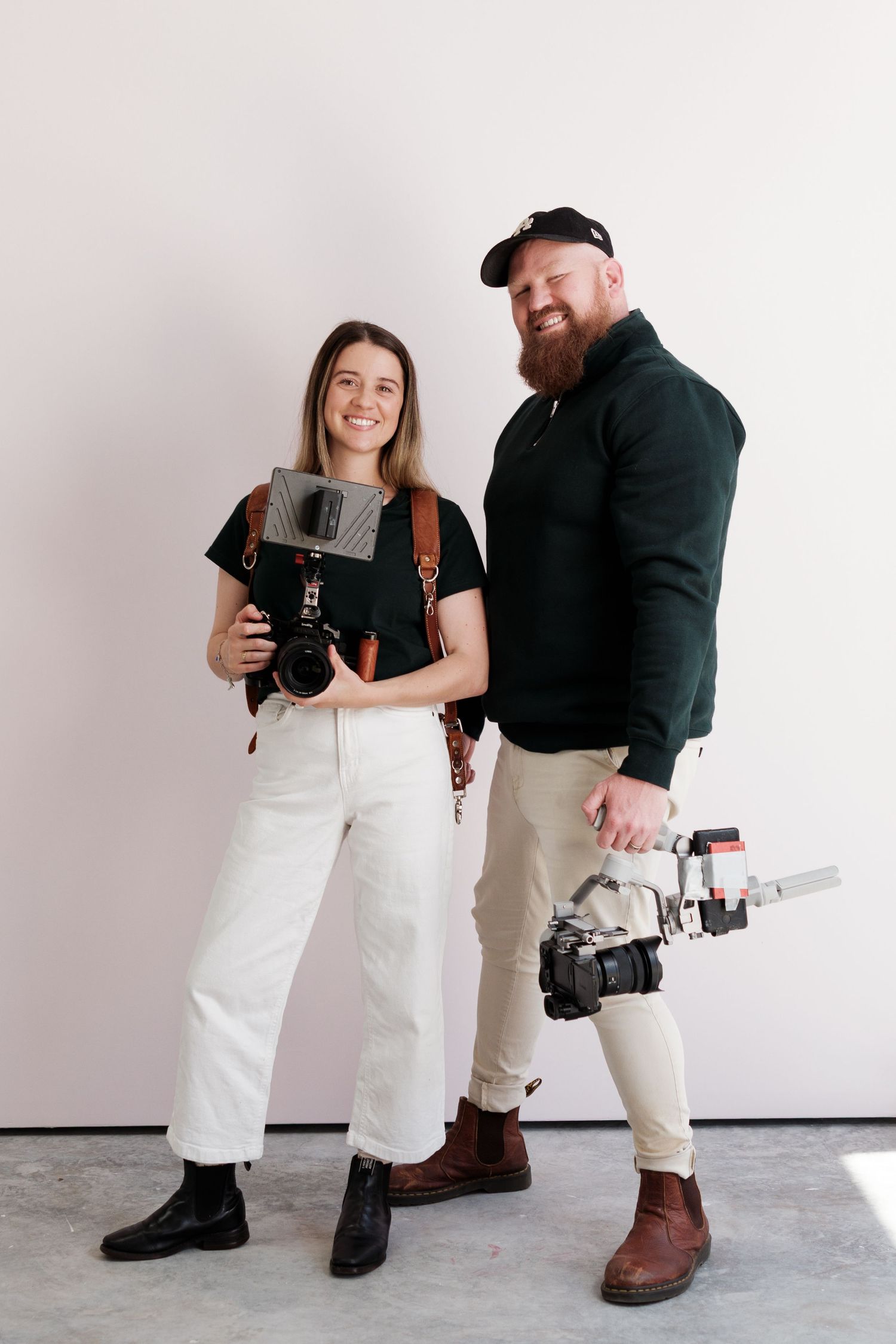 Two photographers stand together holding professional cameras while wearing casual attire against a light background.