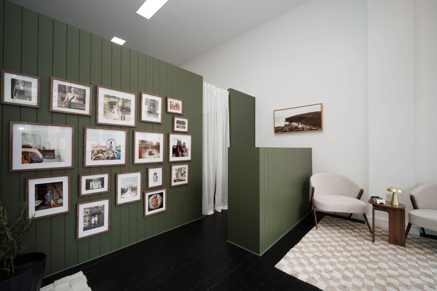 Modern interior with dark wood floors, green shiplap gallery wall displaying framed photos, and a cozy white armchair in the corner.