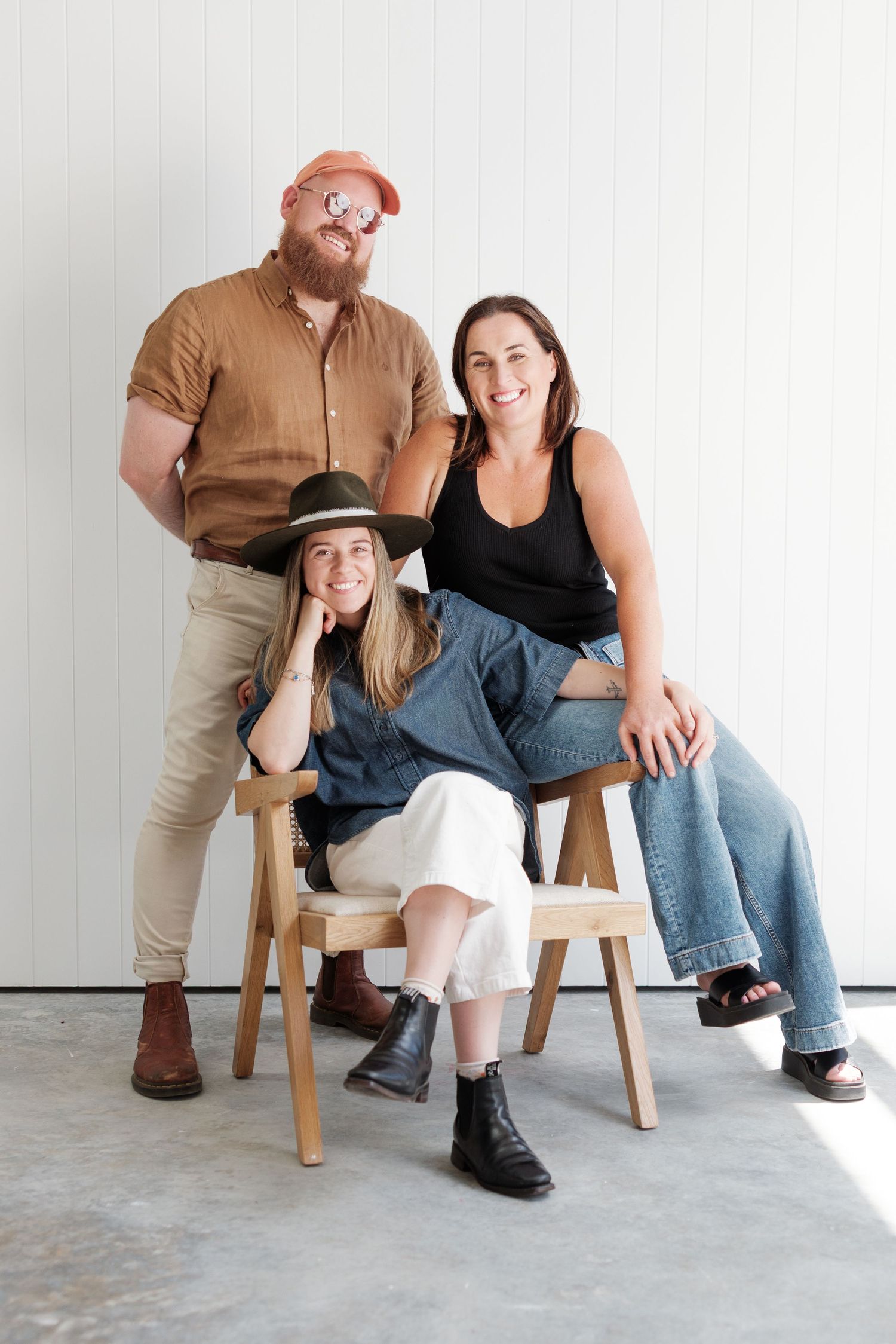 Two people sit casually on wooden chairs against a white wall in a minimalist studio setting.