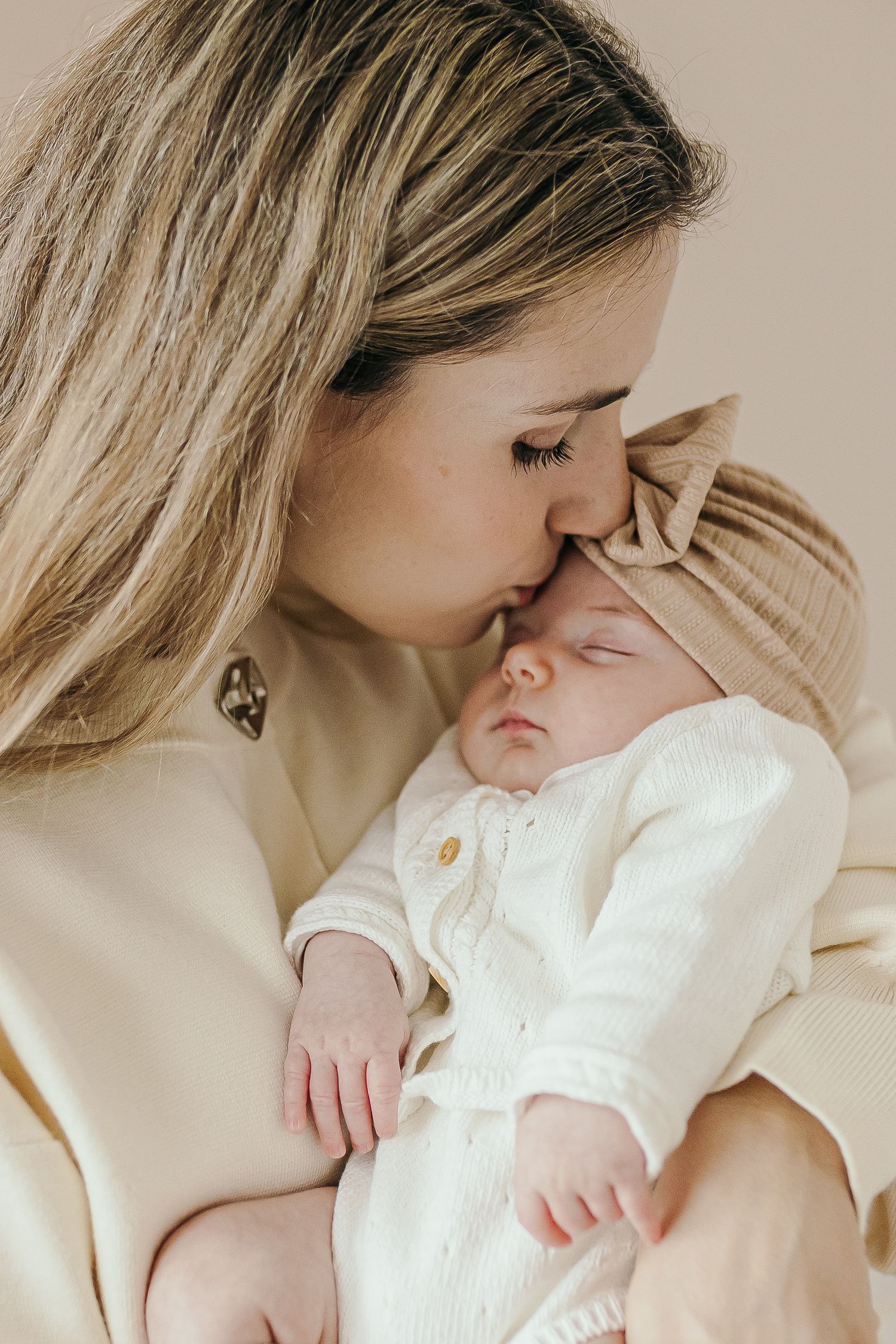 A mother tenderly kisses her sleeping newborn baby dressed in white, wearing a beige knot turban hat.