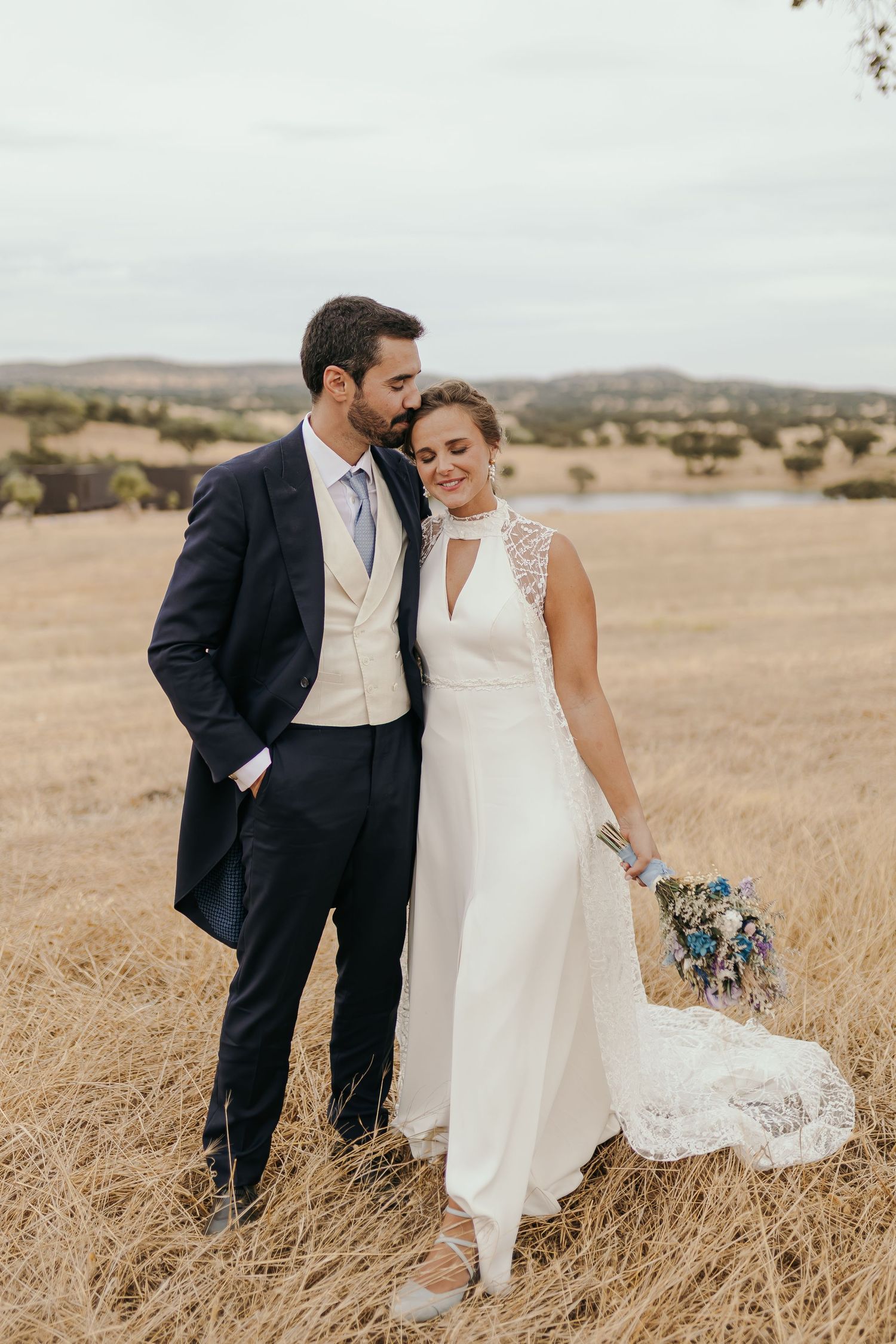 Bride and groom pose in golden field, she wears white lace gown holding bouquet, he wears navy suit.