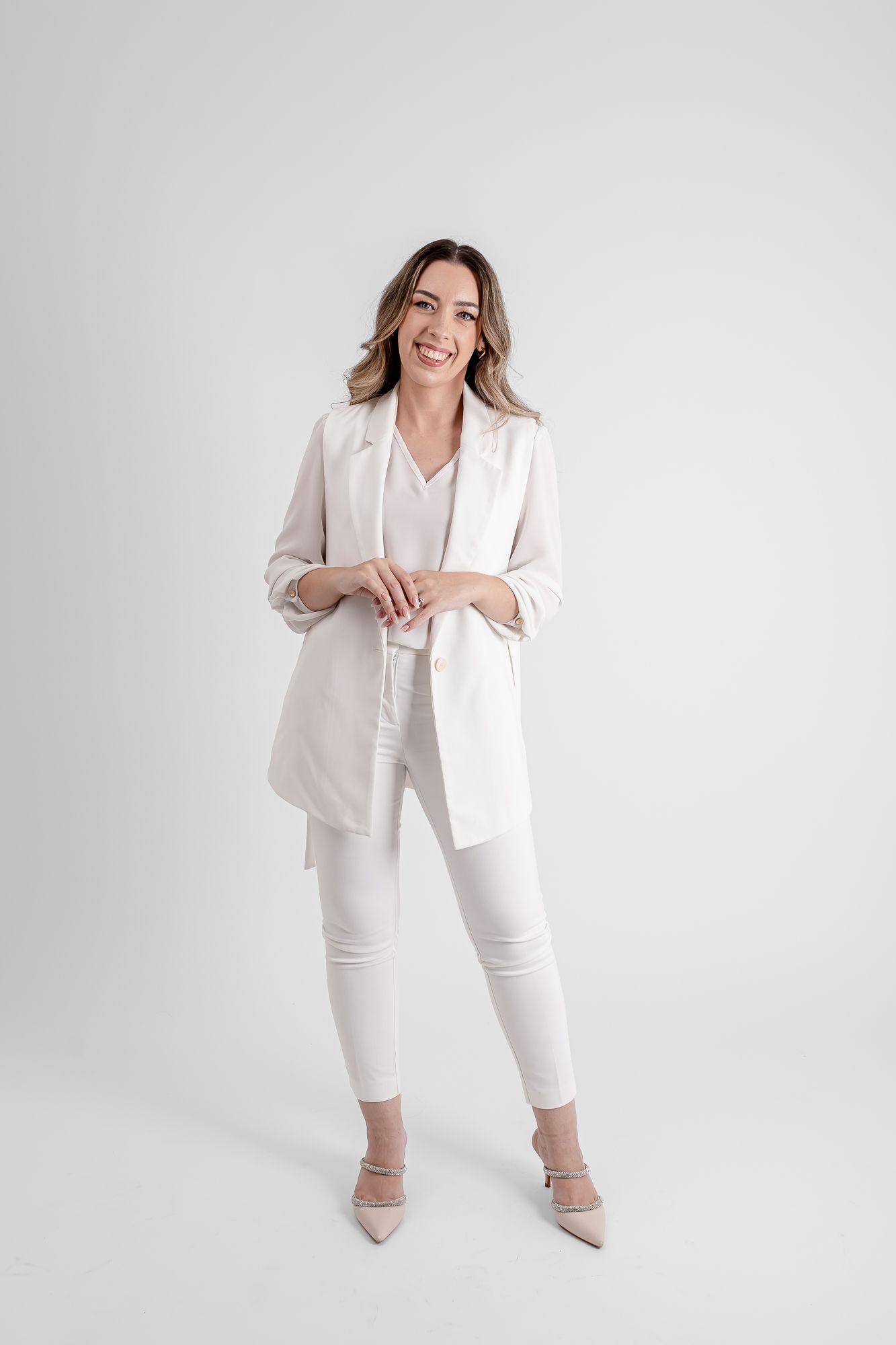 Smiling woman in white blazer, pants, and heels posing confidently against a light gray background.