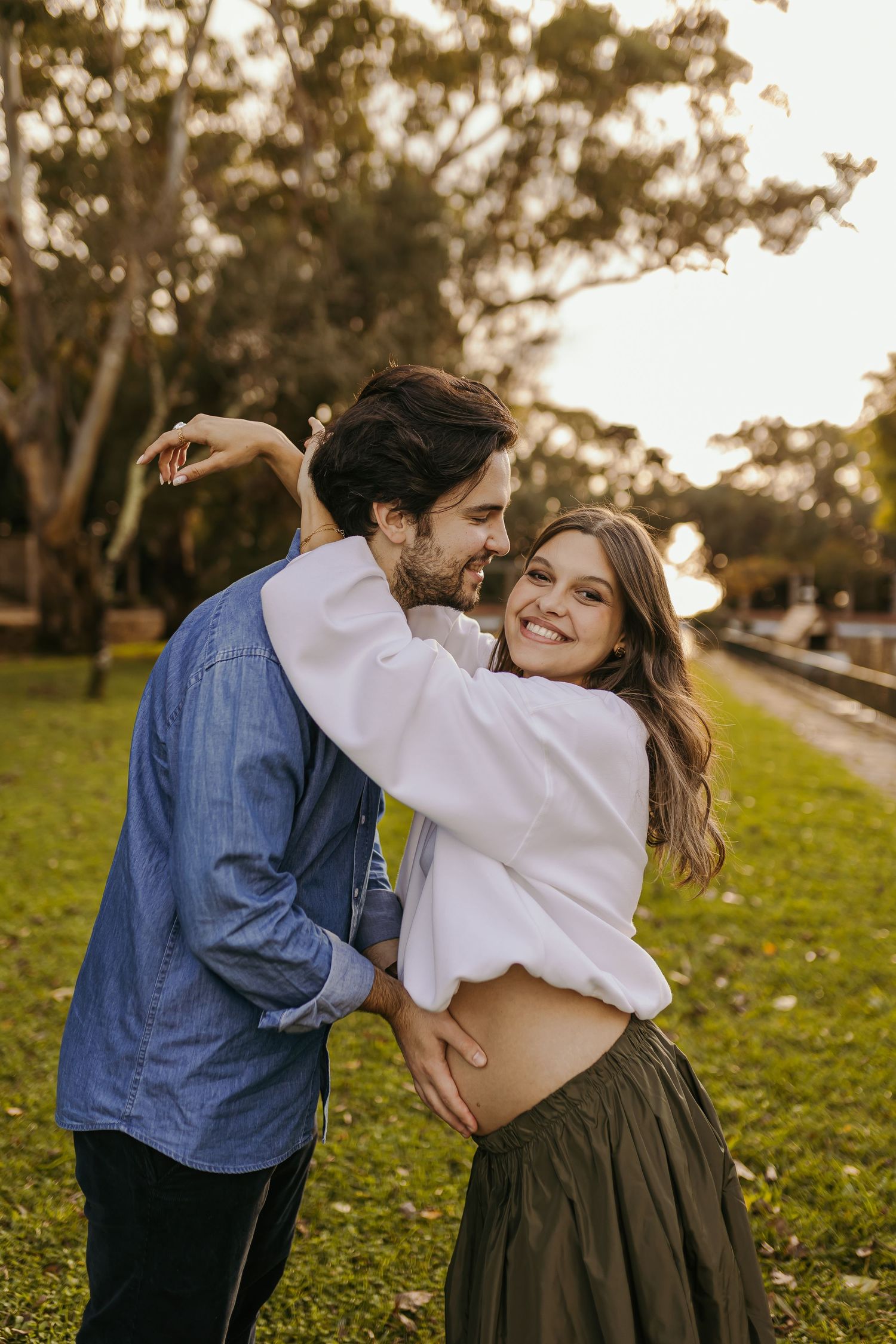 Smiling pregnant couple dancing outdoors in a park at golden hour, woman in white top and green skirt.
