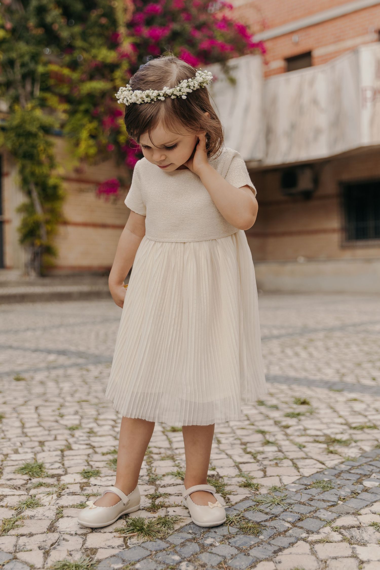 Young flower girl in ivory pleated dress and floral crown standing on cobblestone street.