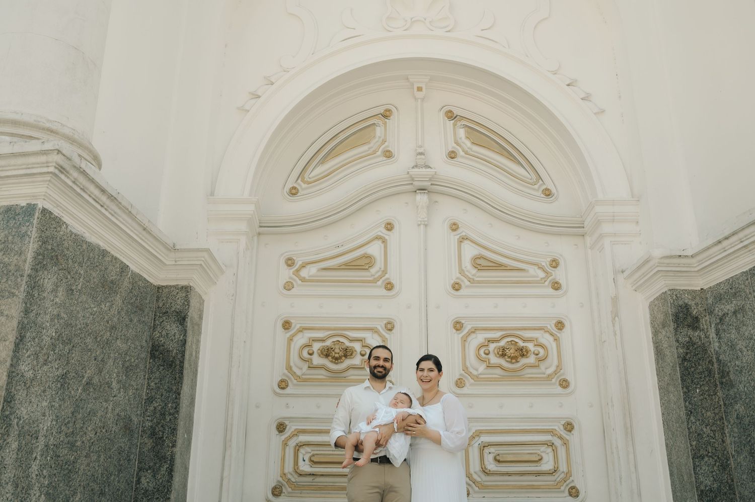 A couple stands in front of an ornate white baroque doorway with green marble columns.