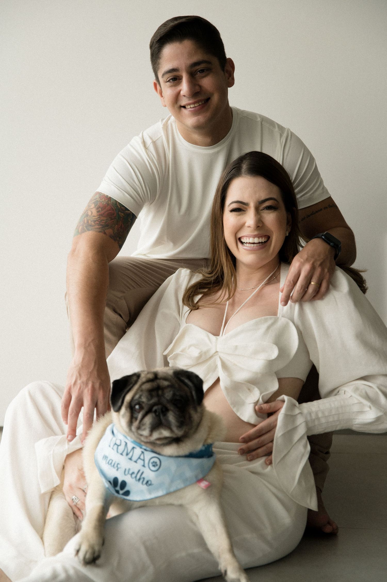 A couple in white clothing poses with their pug dog wearing a blue bandana in a studio portrait.