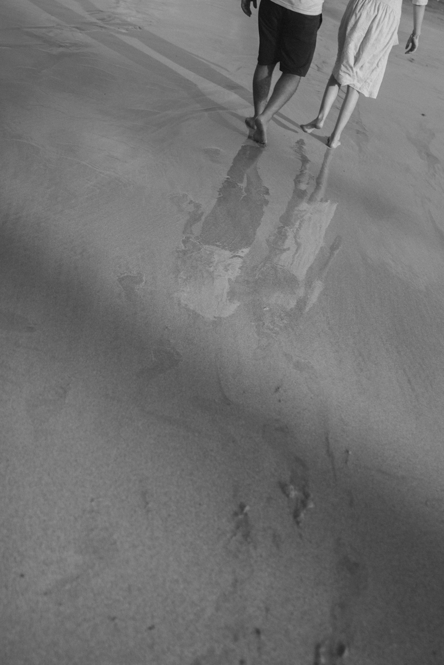 Black and white image of footprints and shadows on wet sand at the beach.