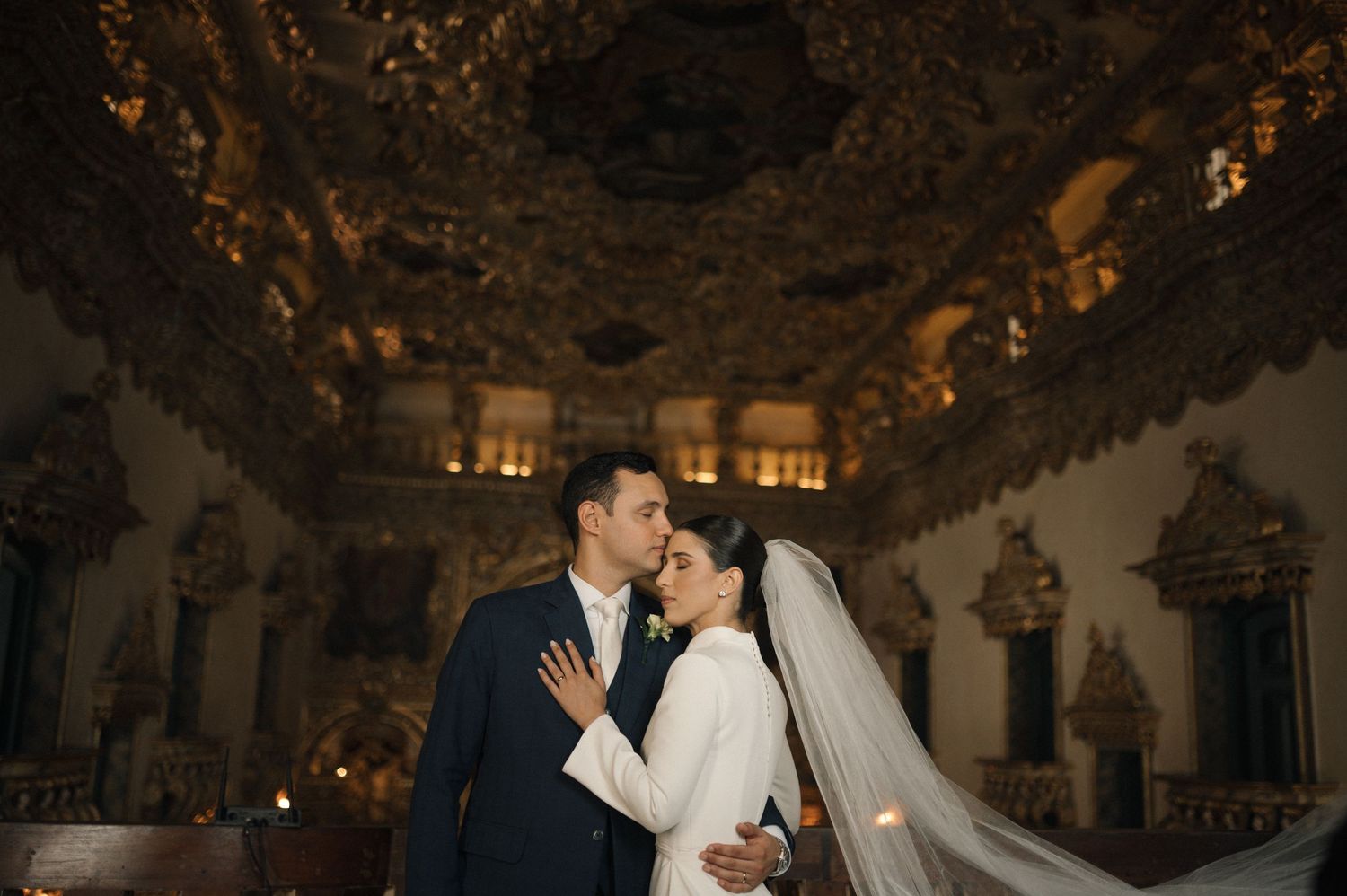 Bride and groom share a romantic moment under ornate golden baroque ceiling in an elegant historic venue.