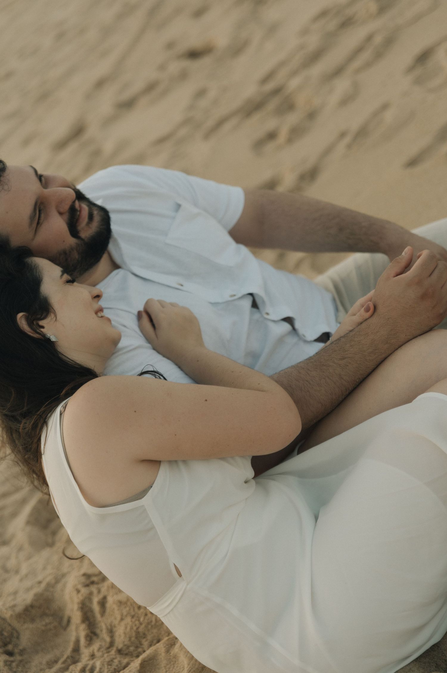 A couple in white clothing lying together on golden sand.