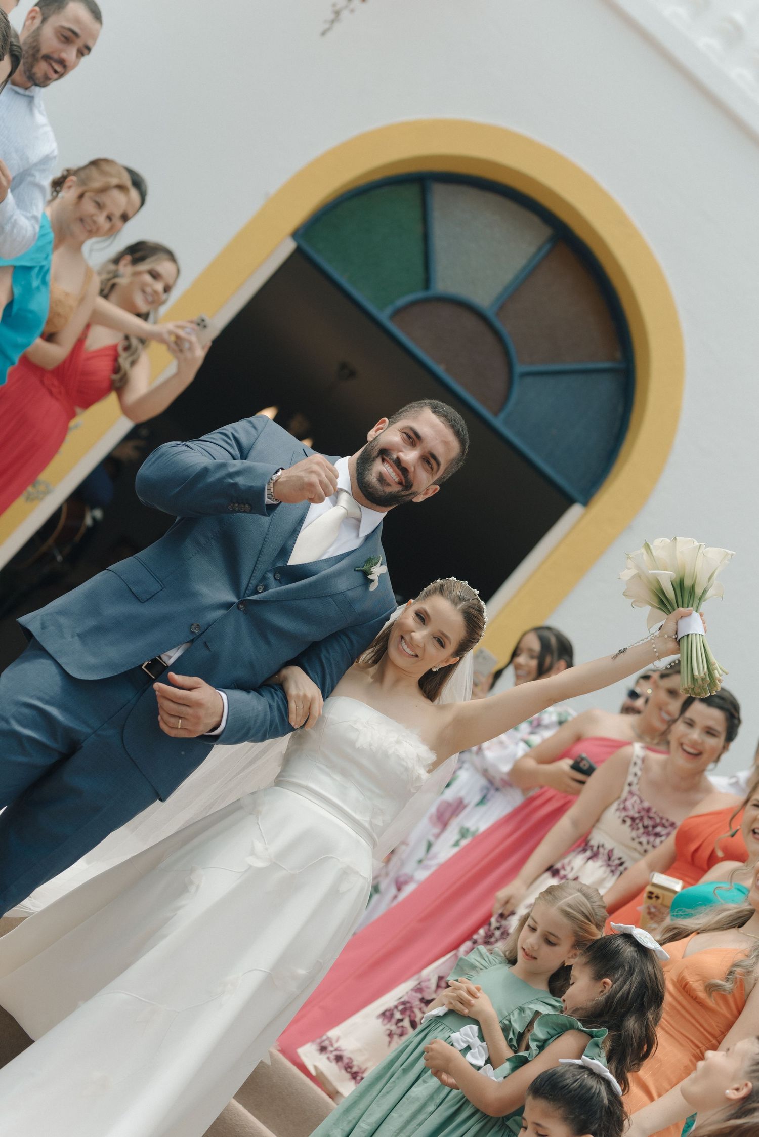 Joyful wedding ceremony exit with guests in bright dresses cheering beneath a decorative archway.