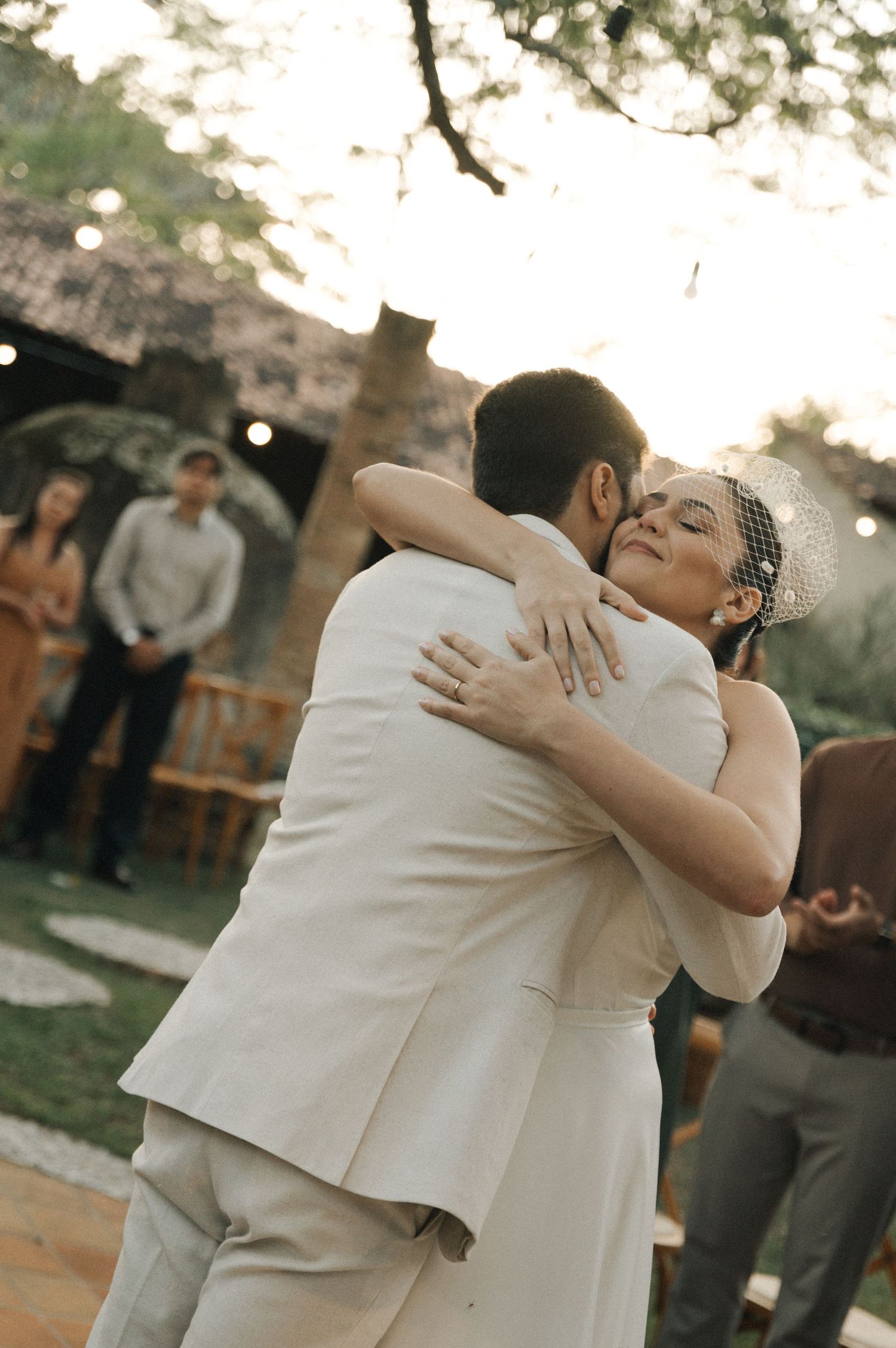 Two people share an emotional embrace during an outdoor wedding celebration at sunset.