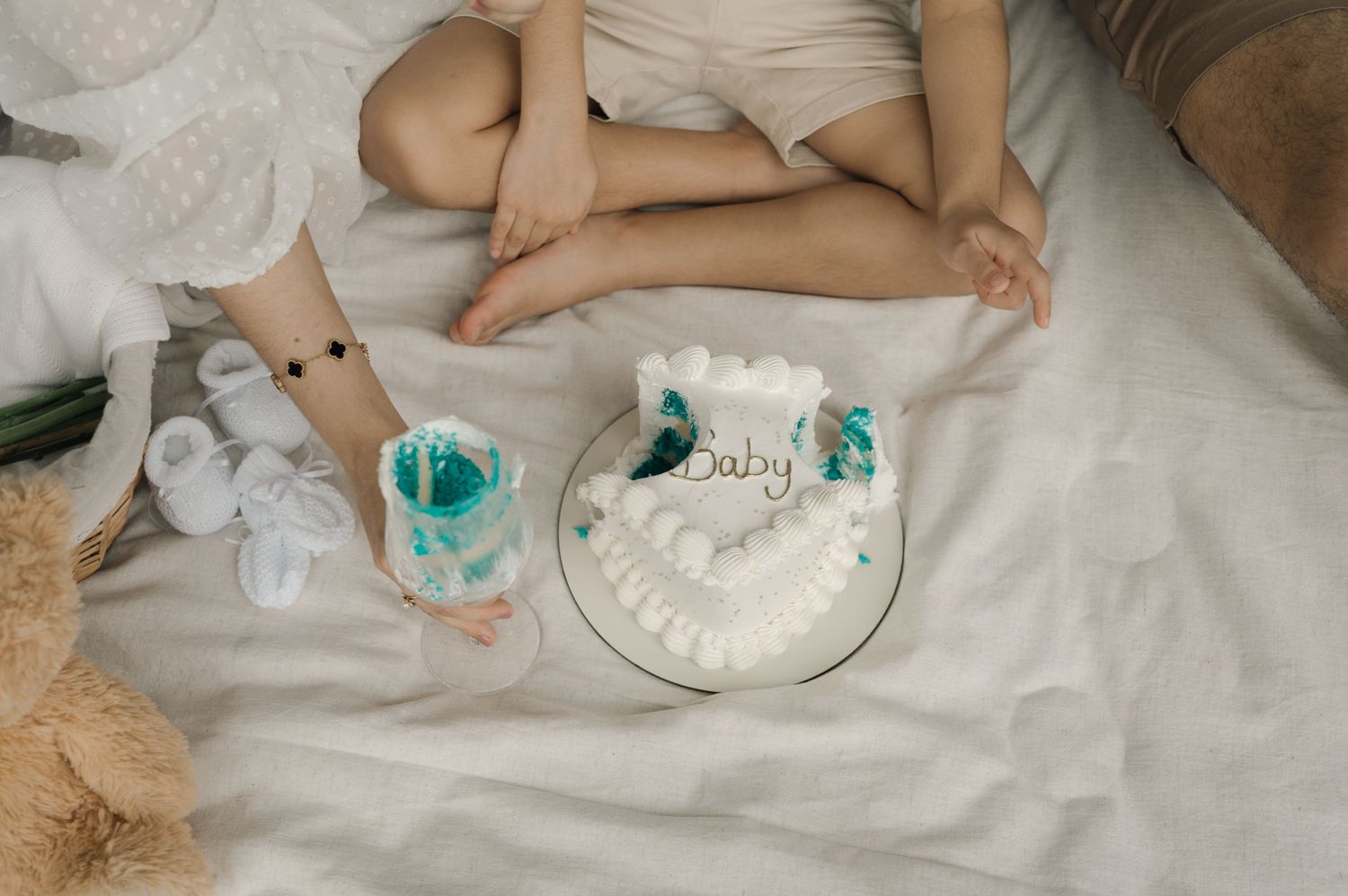 Two children sit on white bedding beside a baby shower cake decorated with teal accents and tiny booties.