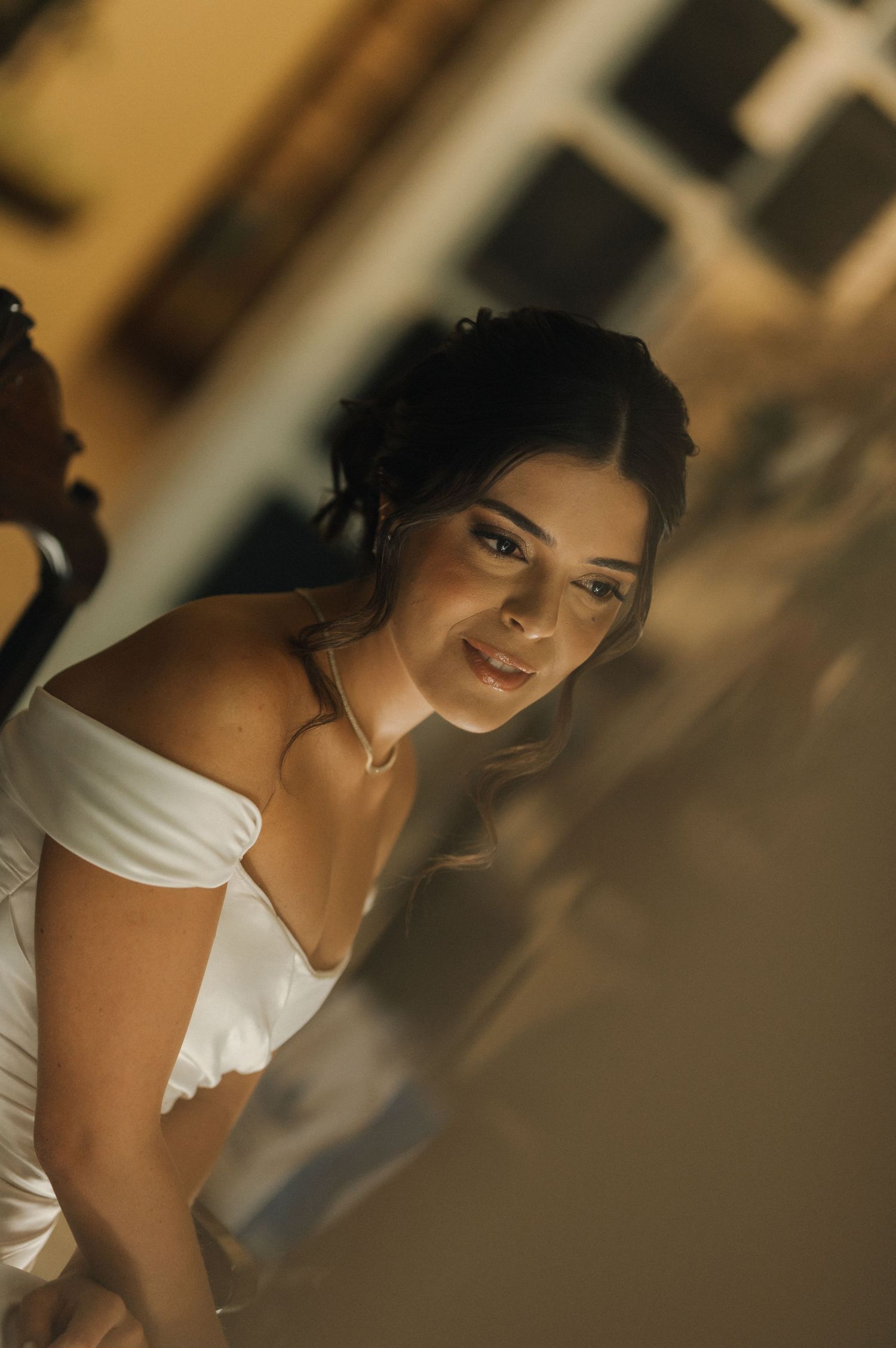 A bride in an off-shoulder white gown smiles warmly in warm ambient lighting.