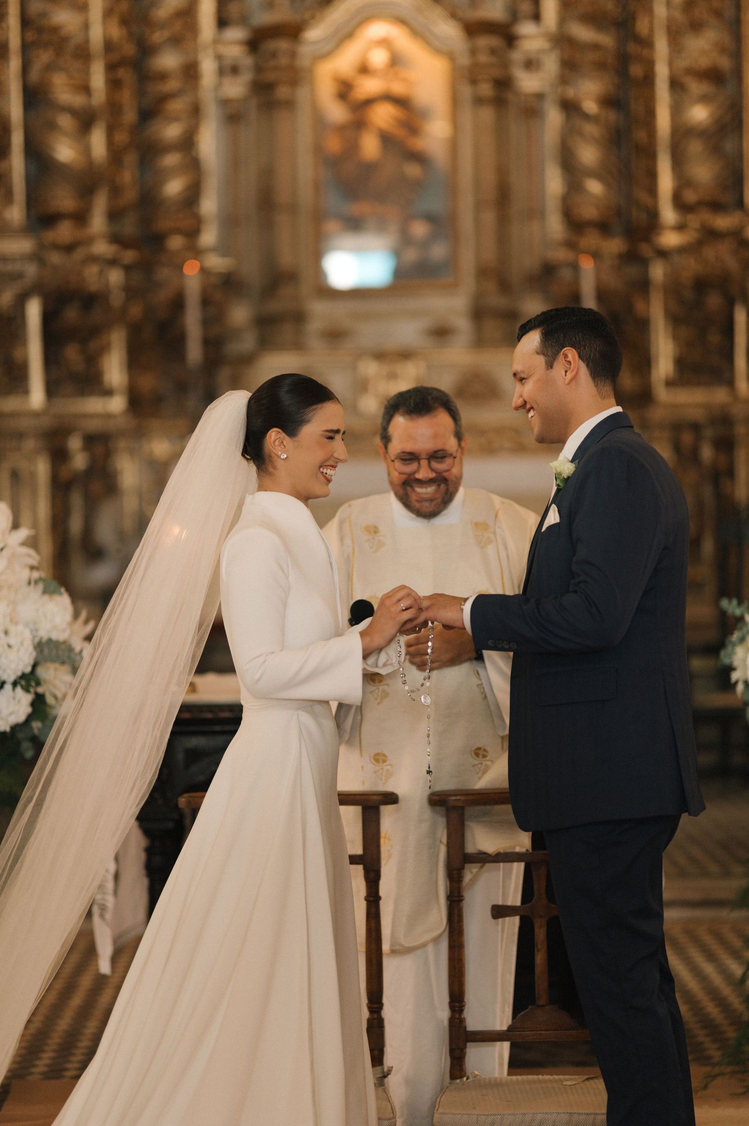Bride and groom exchange rings at church altar while smiling, officiated by priest in ornate golden church.