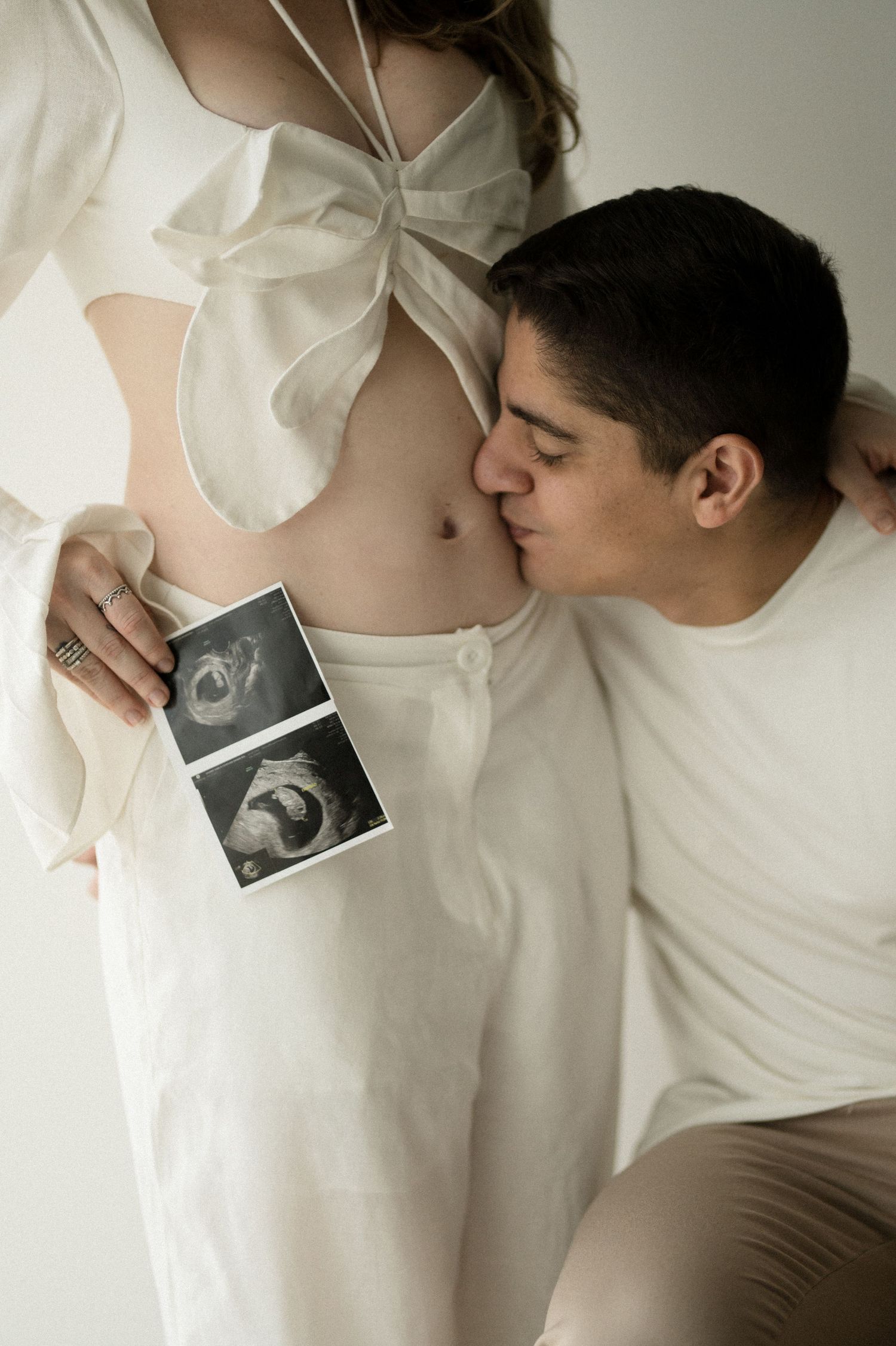 A couple in white clothing holds an ultrasound photo against a pregnant belly in a series of tender maternity photos.