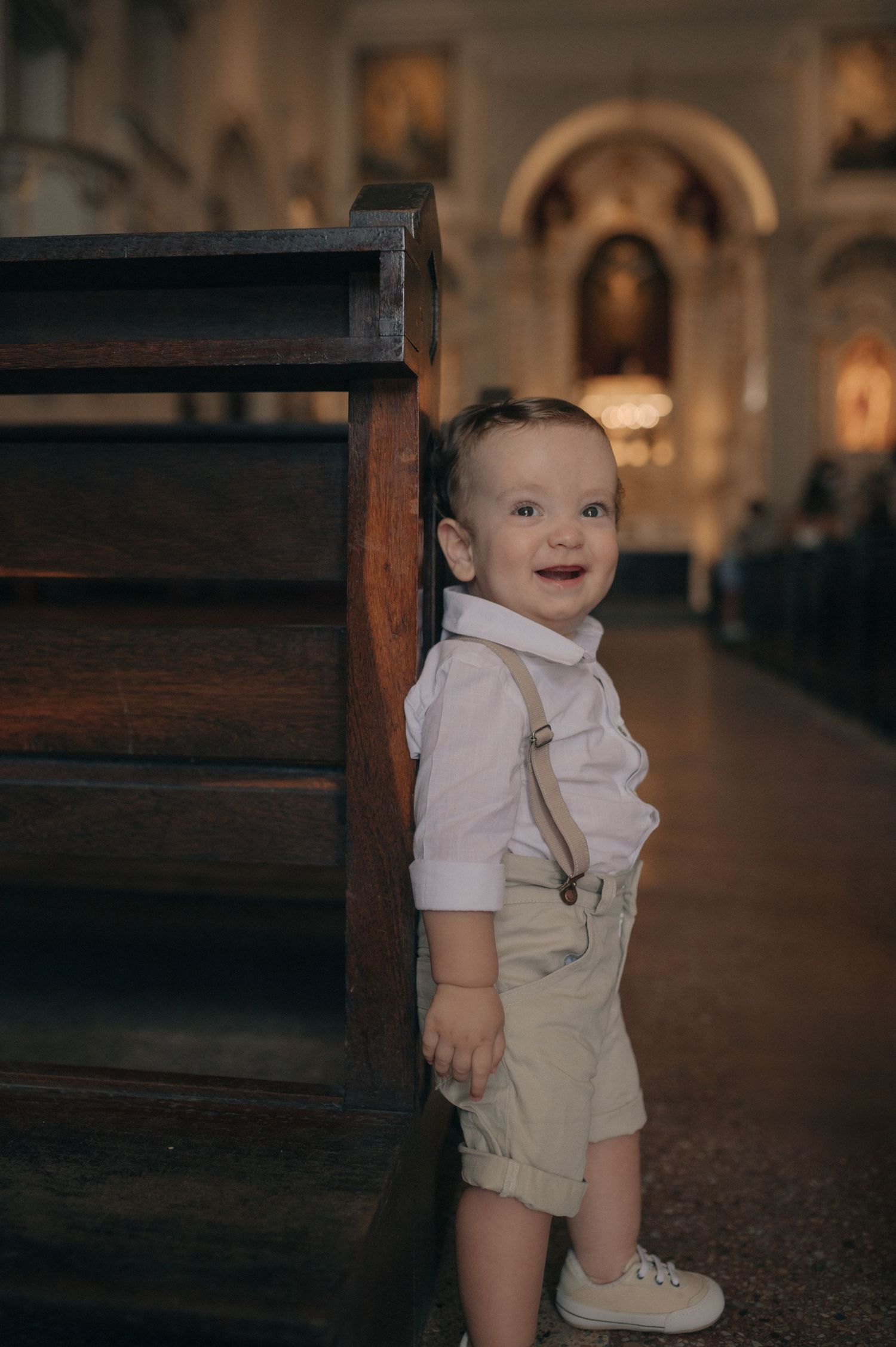 A small child in white overalls and shirt sits on wooden church pews in a dimly lit cathedral interior.
