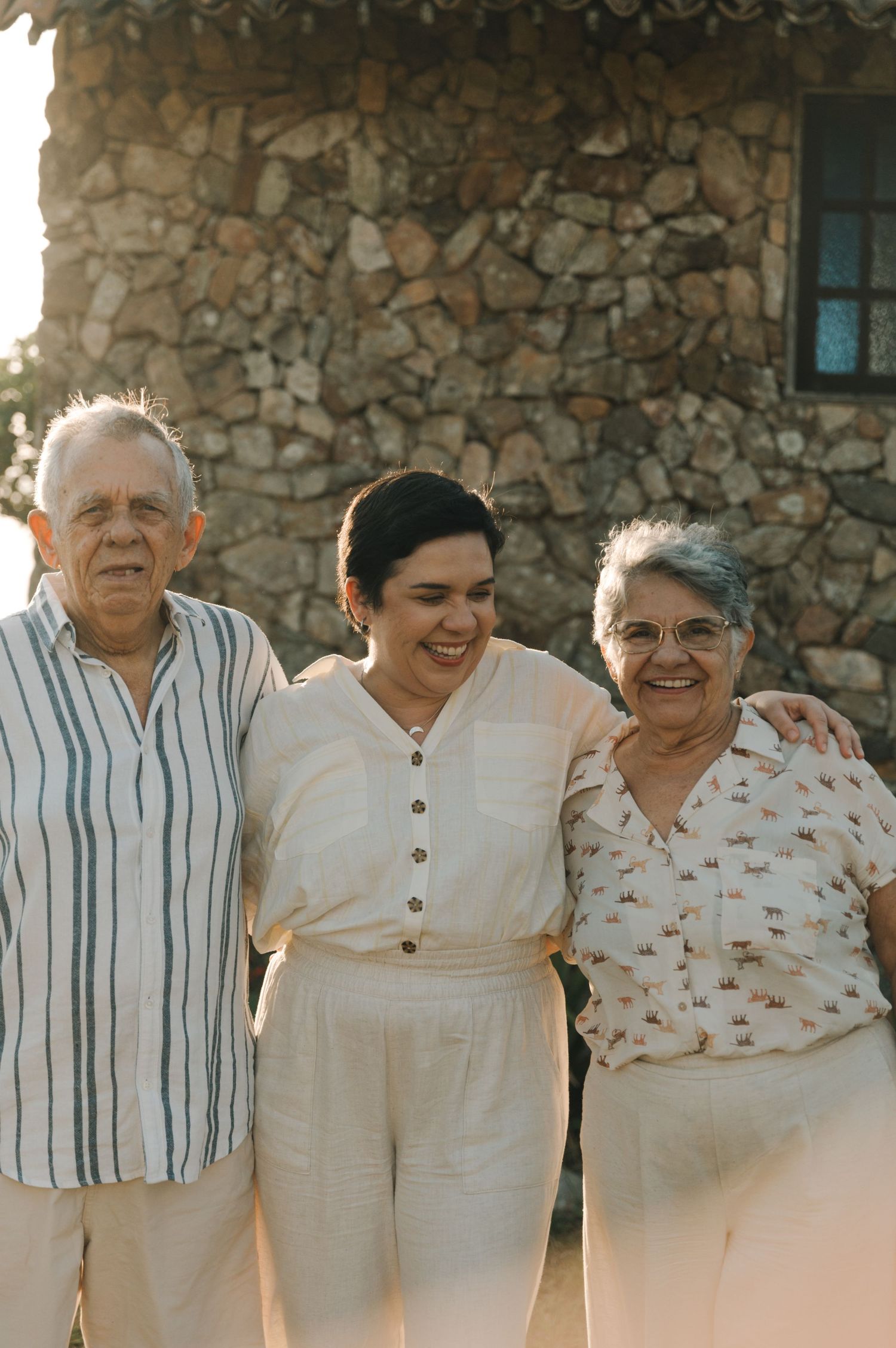 A group of elderly people wearing light colored clothing standing together against a stone wall.
