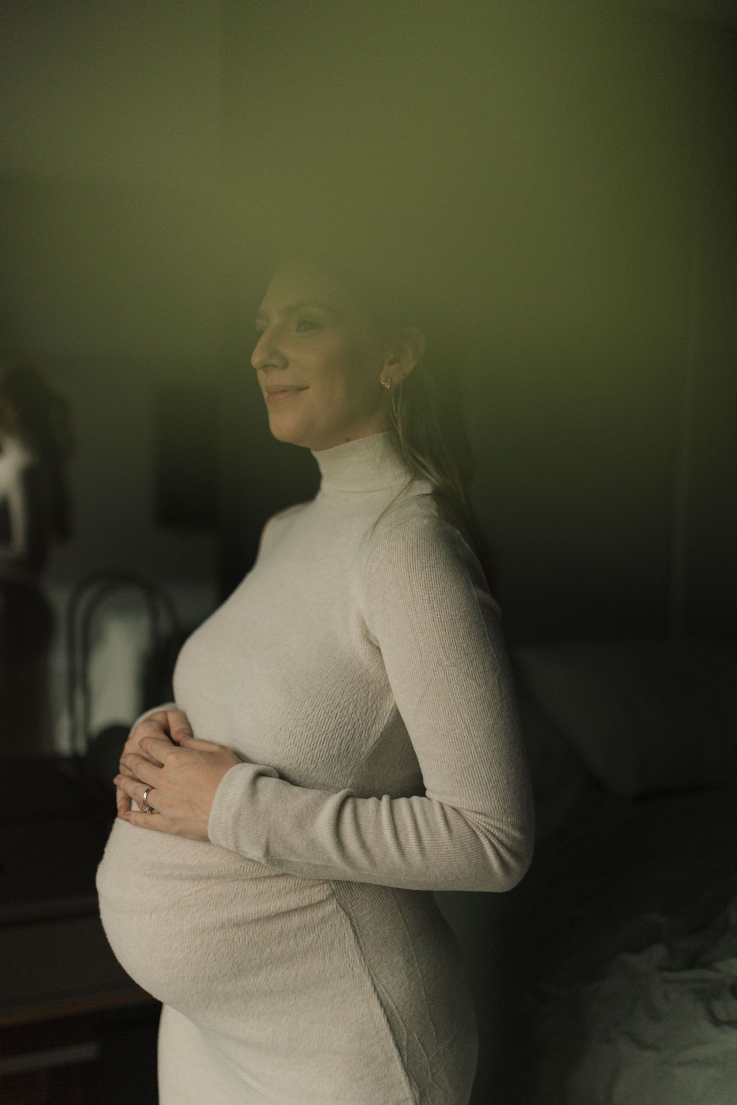 Expectant mother in cream turtleneck sweater dress poses near window with gentle smile.