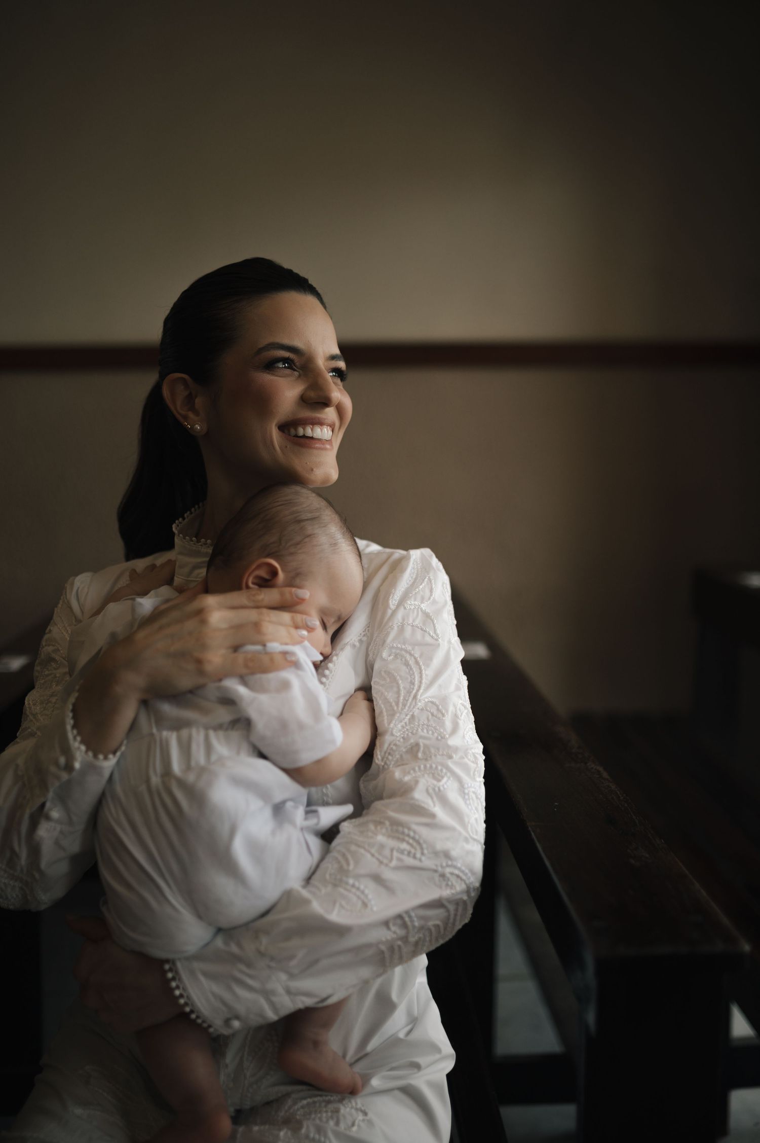 Smiling woman in white lace dress holding a sleeping baby in a warmly lit church setting.